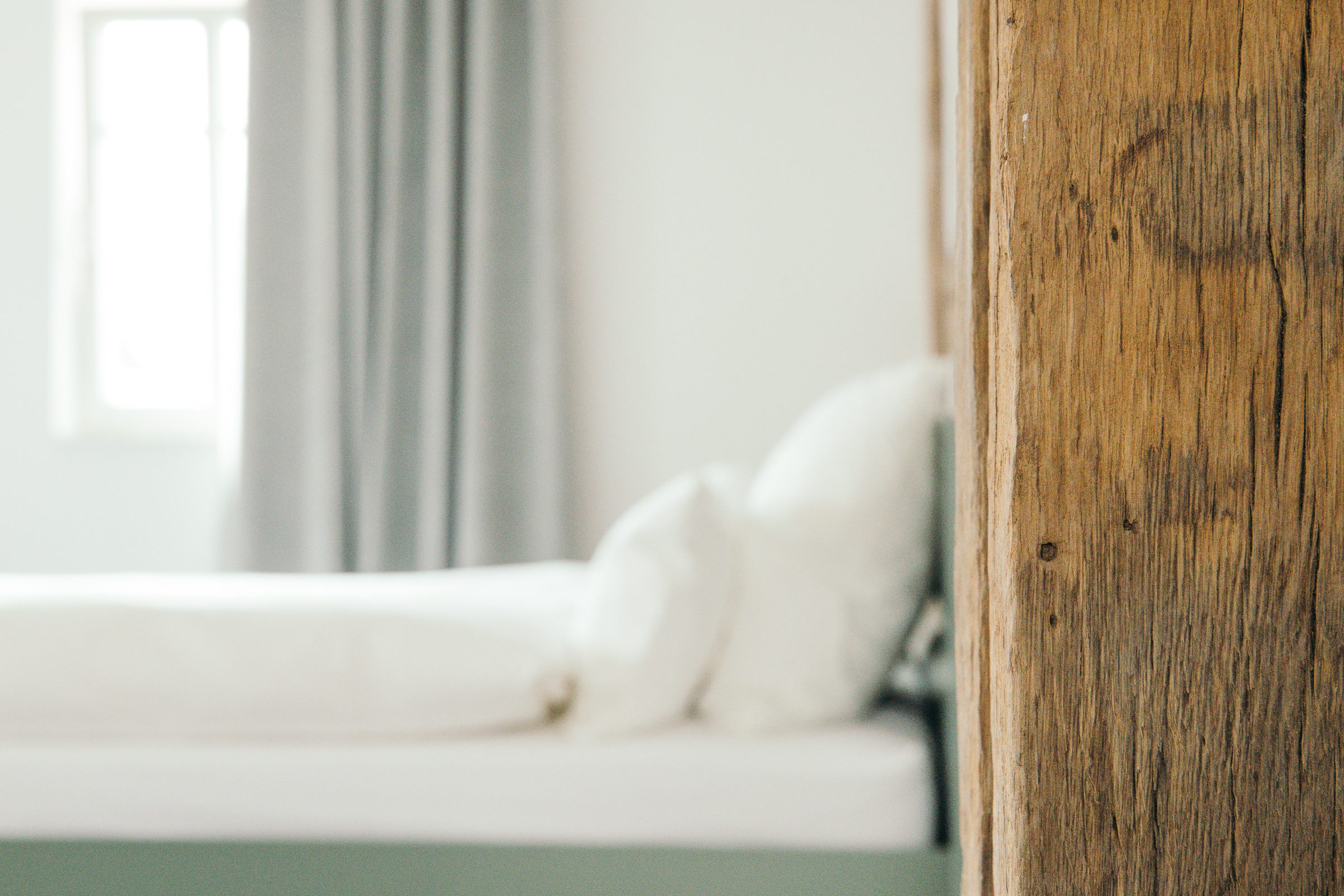 Bett mit weißen Kissen vor einem hellen Fenster, rechts im Vordergrund ein alter Holzbalken
