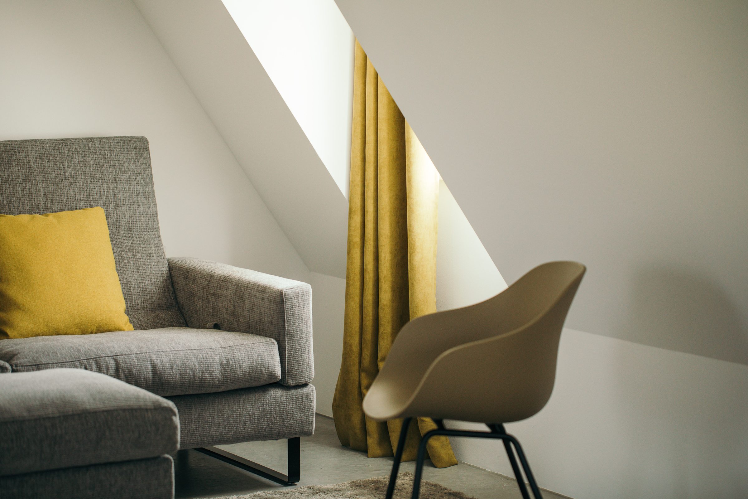 Graues Sofa mit gelbem Kissen und ein Sessel unter der Dachschräge, daneben goldgelbe Vorhänge