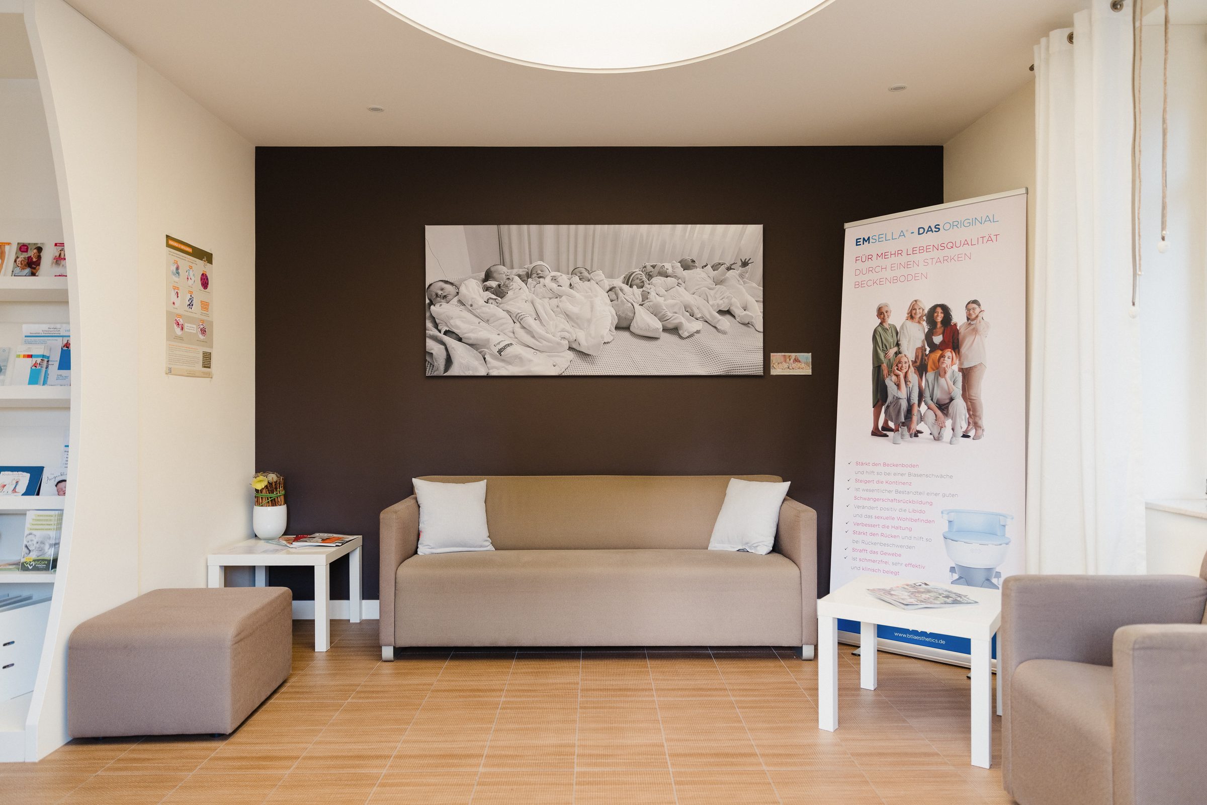 Wartebereich der Praxis Dr. Meyer-Wilmes mit beigem Sofa, Sesseln und einem großen Schwarzweißfoto von Babys an der dunklen Wand