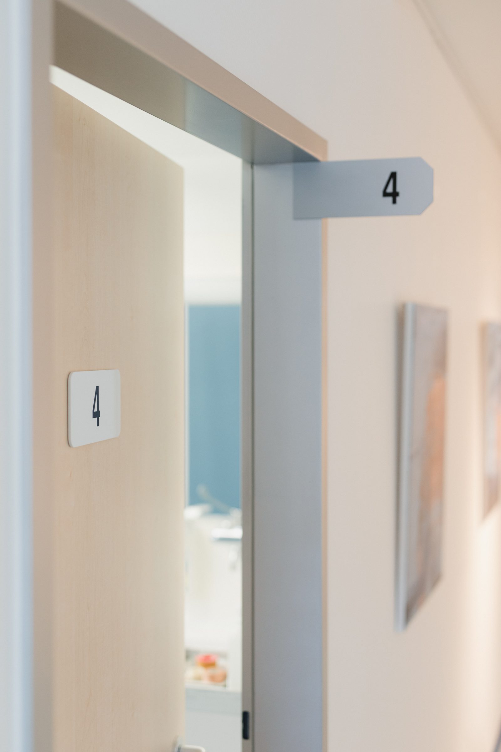 Geöffnete Tür mit der Nummer 4 zu einem Behandlungsraum, daneben Bilder an der Flurwand