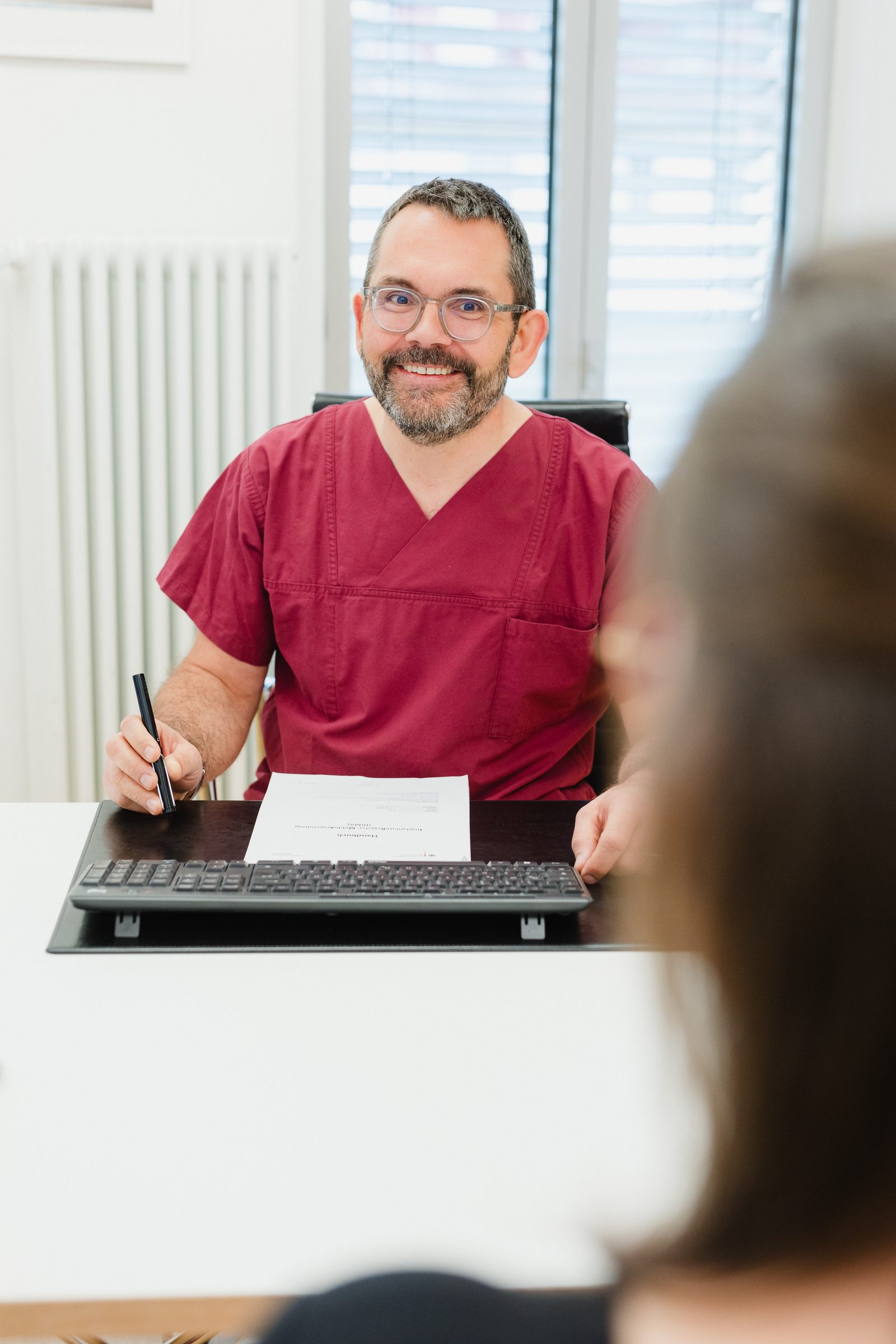 Arzt in weinroter Praxiskleidung sitzt lächelnd mit Stift und Unterlagen am Schreibtisch, vorne unscharf eine Person von hinten