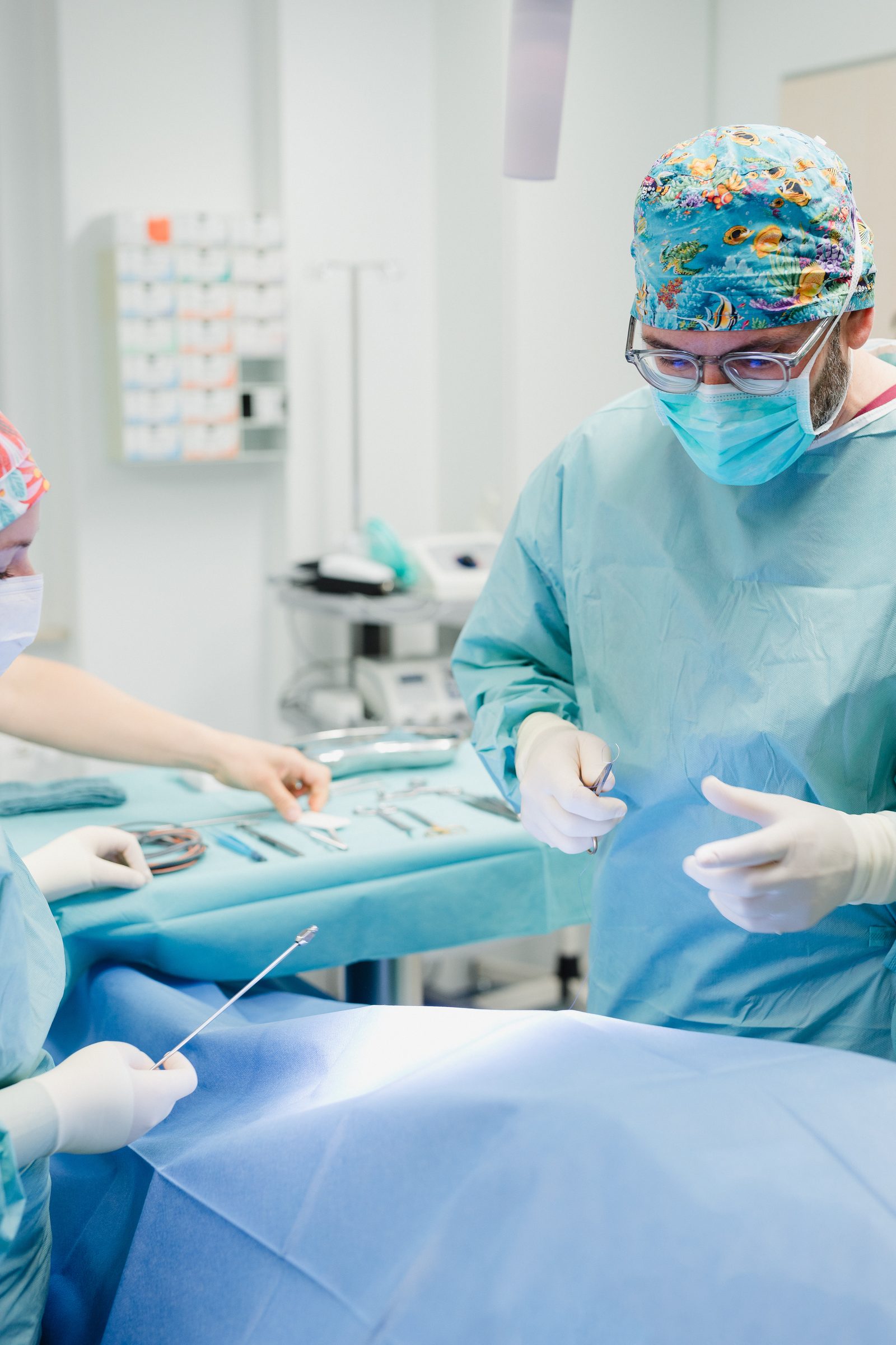 Arzt mit bunter OP-Haube, Mundschutz und Kittel bei einer Operation, links das OP-Team am Instrumententisch