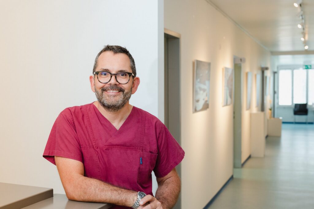 Arzt in weinroter Praxiskleidung lehnt lächelnd an einem Tresen, dahinter ein heller Flur mit Bildern an den Wänden