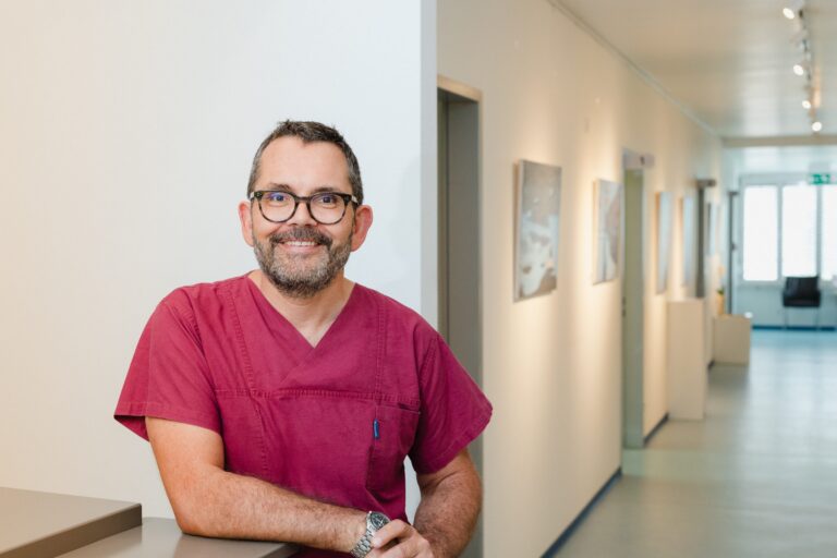 Arzt in weinroter Praxiskleidung lehnt lächelnd an einem Tresen, dahinter ein heller Flur mit Bildern an den Wänden
