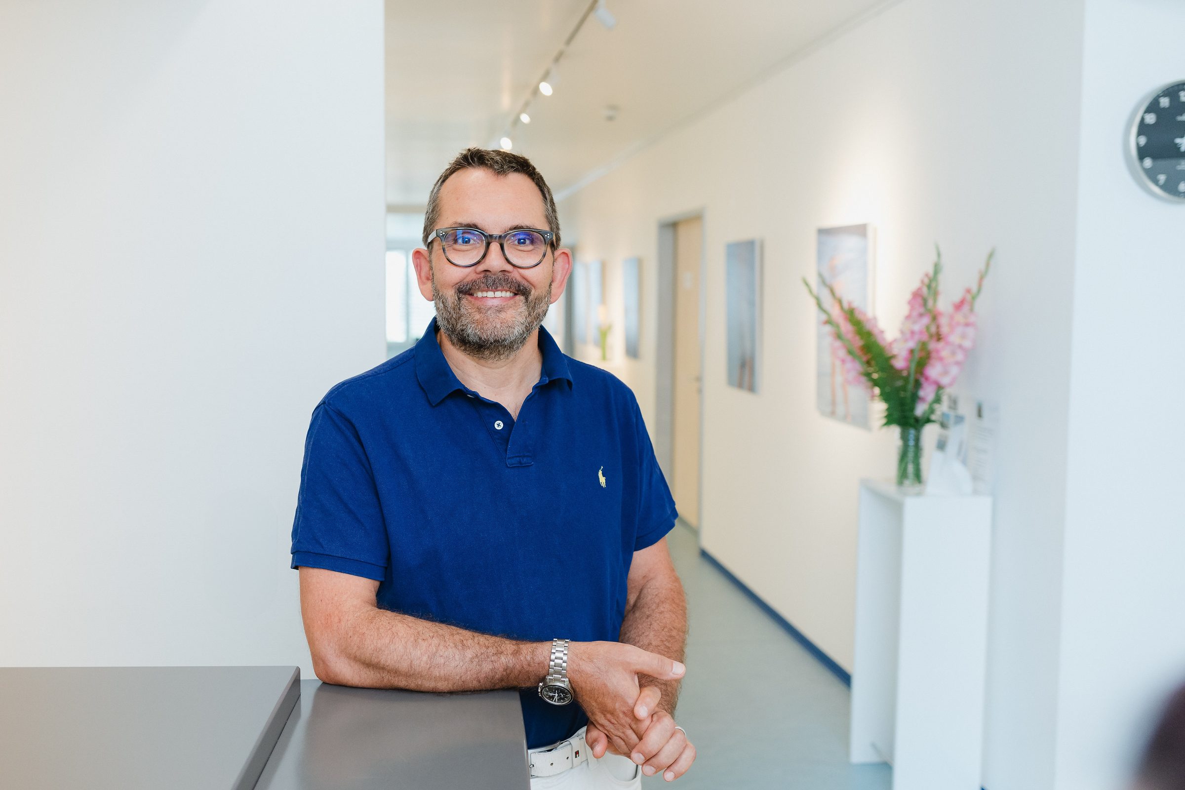Arzt im blauen Poloshirt lehnt lächelnd an einem Tresen im hellen Praxisflur, daneben rosa Blumen in einer Vase