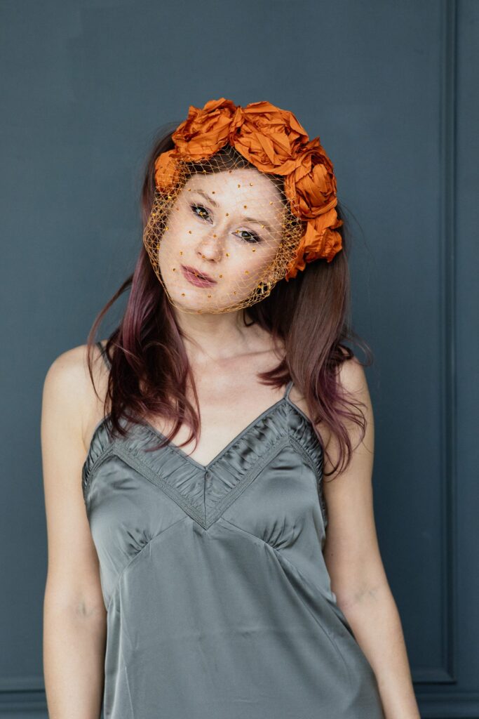 Porträt von Sasha Walker mit orangefarbenem Blütenkopfschmuck und Netzschleier im grauen Satintop vor einer blaugrauen Wand