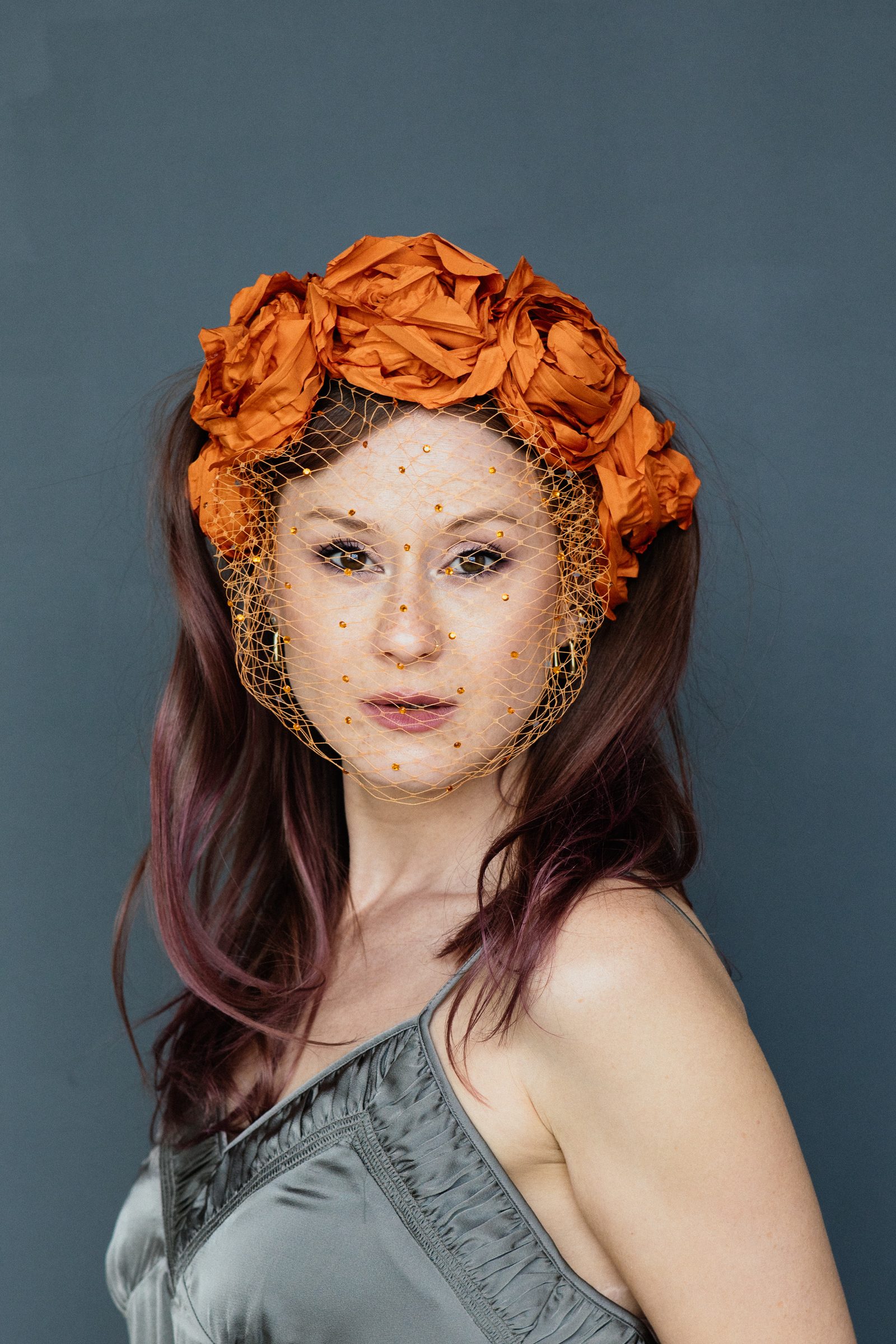 Sasha Walker mit orangefarbenem Blütenkopfschmuck blickt über die Schulter in die Kamera, vor einer blaugrauen Wand