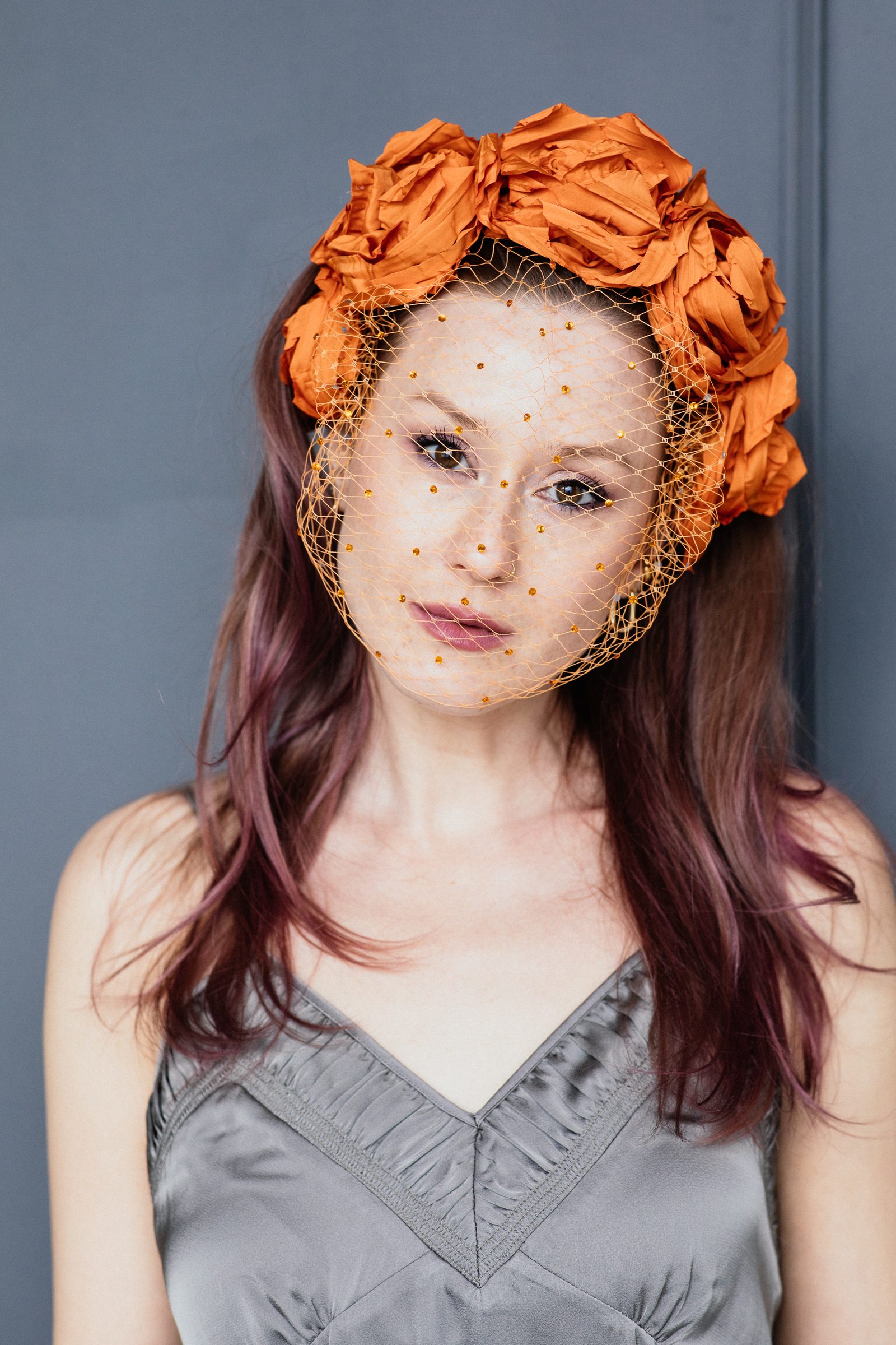 Nahaufnahme von Sasha Walker mit Blütenkopfschmuck und Netzschleier, frontal vor einer blaugrauen Wand