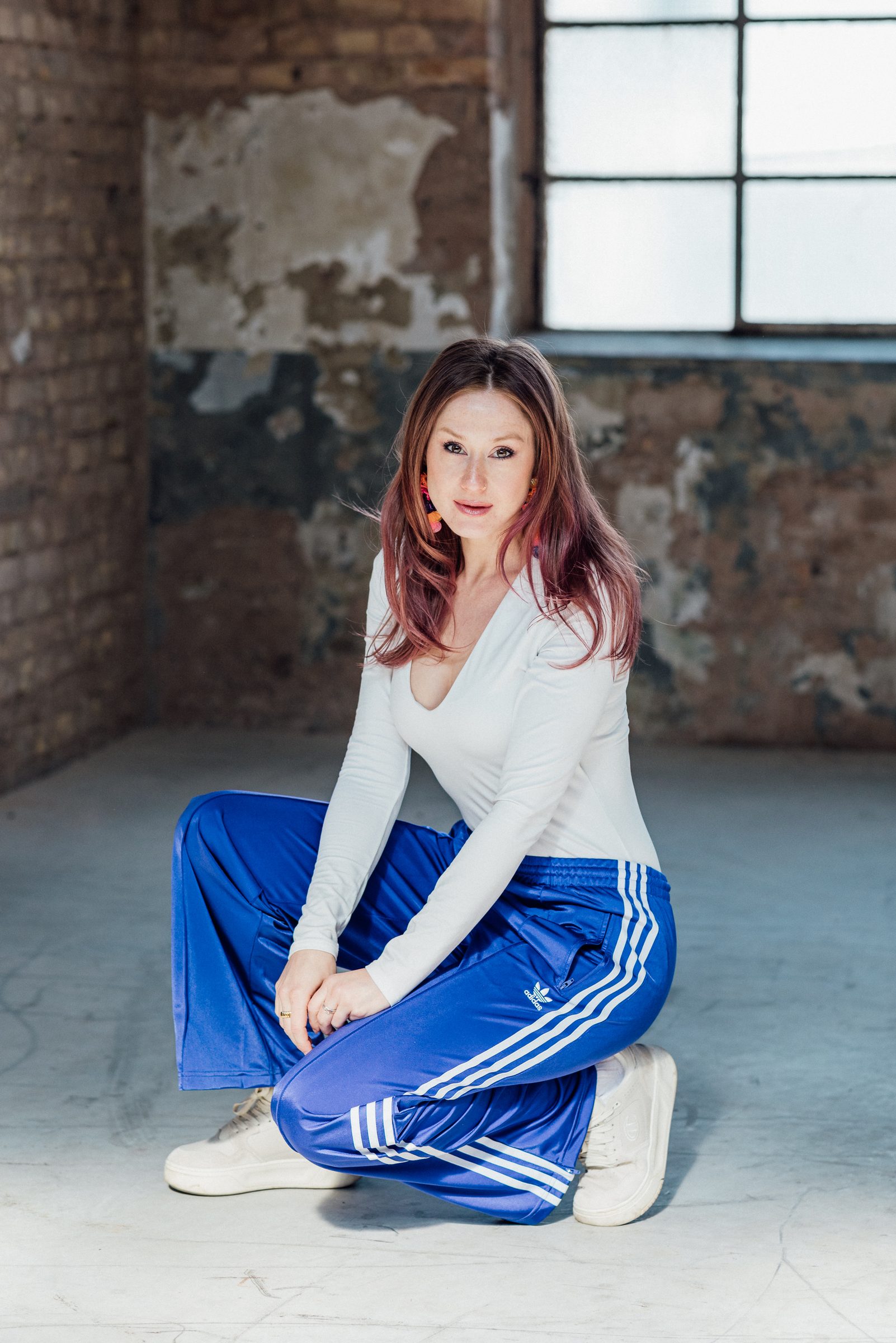 Sasha Walker in weißem Oberteil und blauer Trainingshose hockt in einer Industriehalle mit verwitterter Backsteinwand