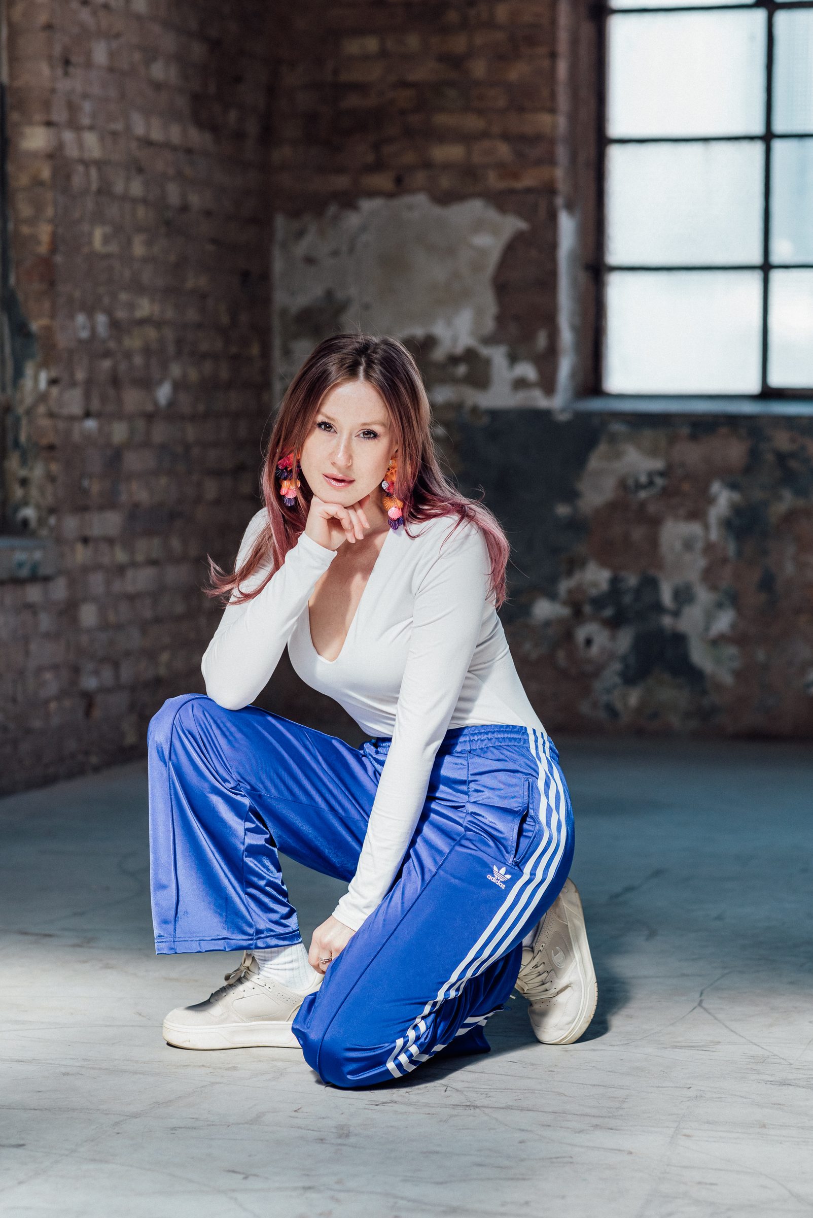 Sasha Walker hockt in der Industriehalle und stützt das Kinn in die Hand, dahinter Backsteinwand und Fenster