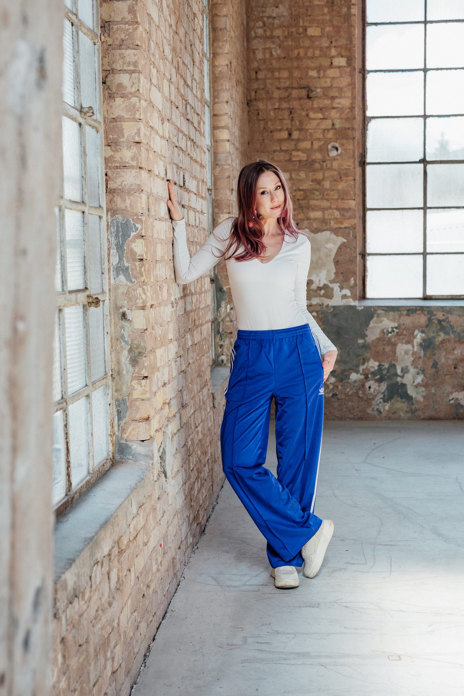 Sasha Walker lehnt in weißem Oberteil und blauer Trainingshose an einer Backsteinwand neben einem Sprossenfenster