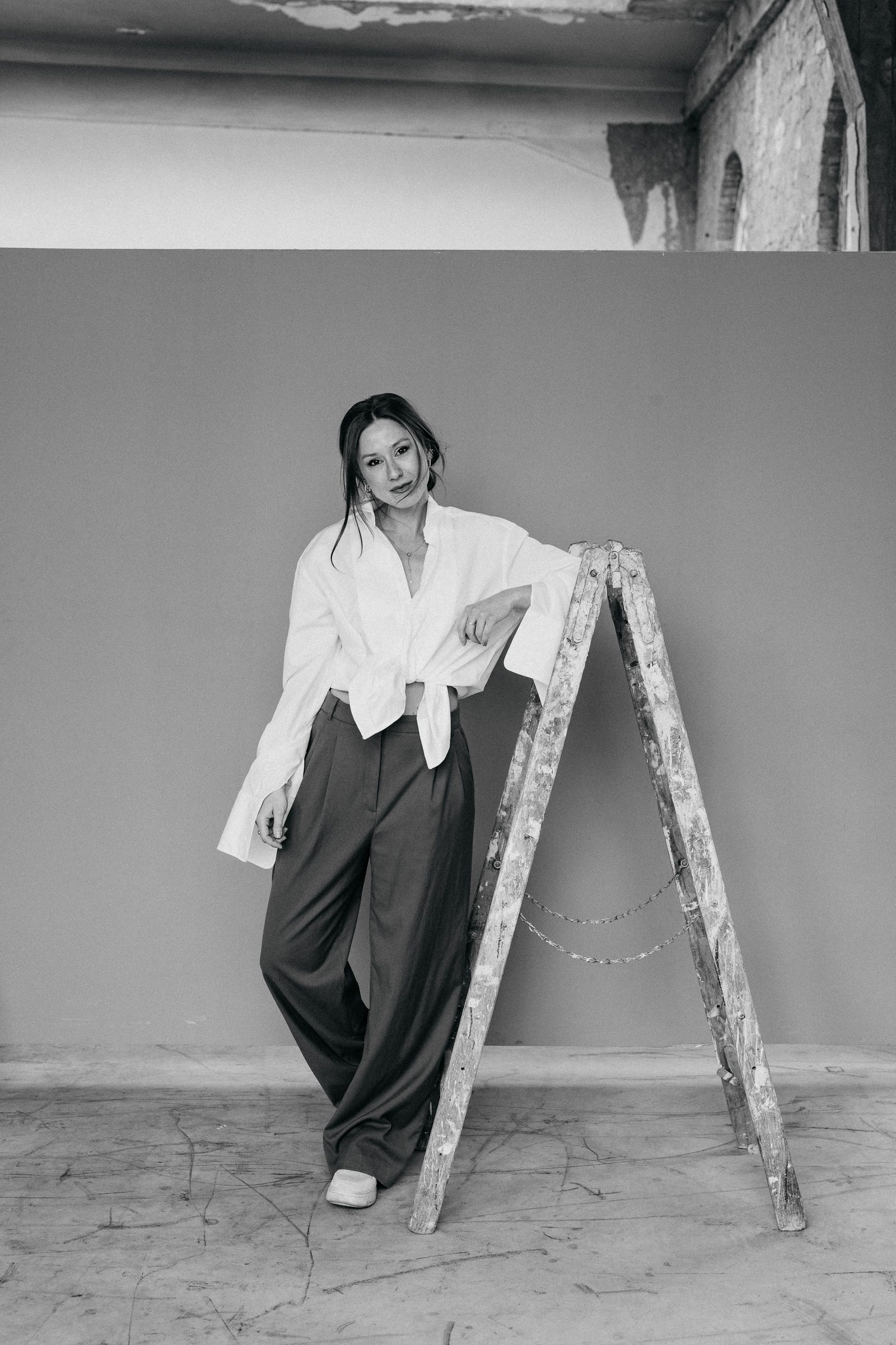 Schwarzweißfoto von Sasha Walker in weißer Bluse und weiter dunkler Hose, an eine Holzleiter gelehnt