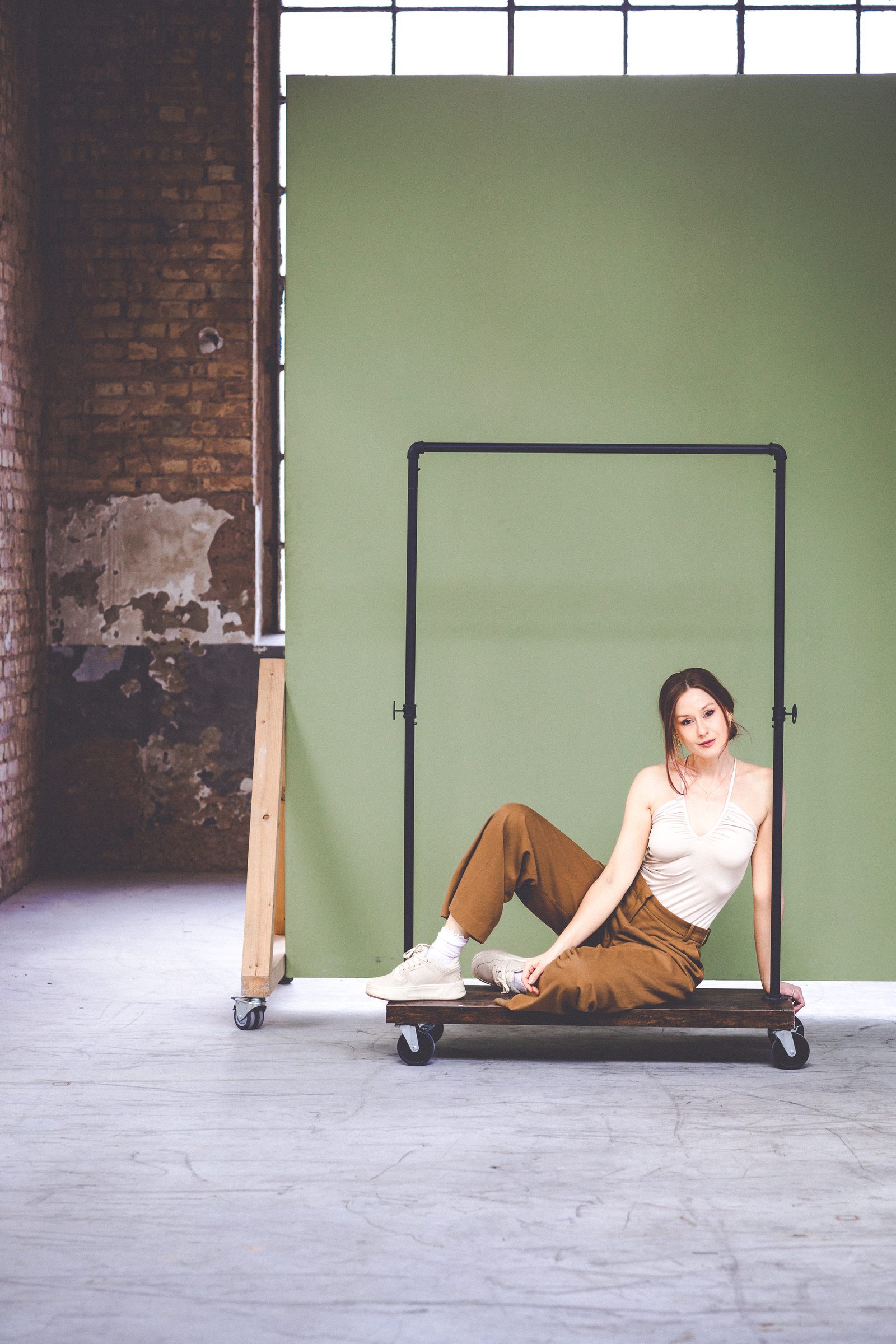 Sasha Walker in weißem Top und brauner Hose sitzt auf einem fahrbaren Kleiderständer im Loft, dahinter Backsteinwand und grüner Fotohintergrund