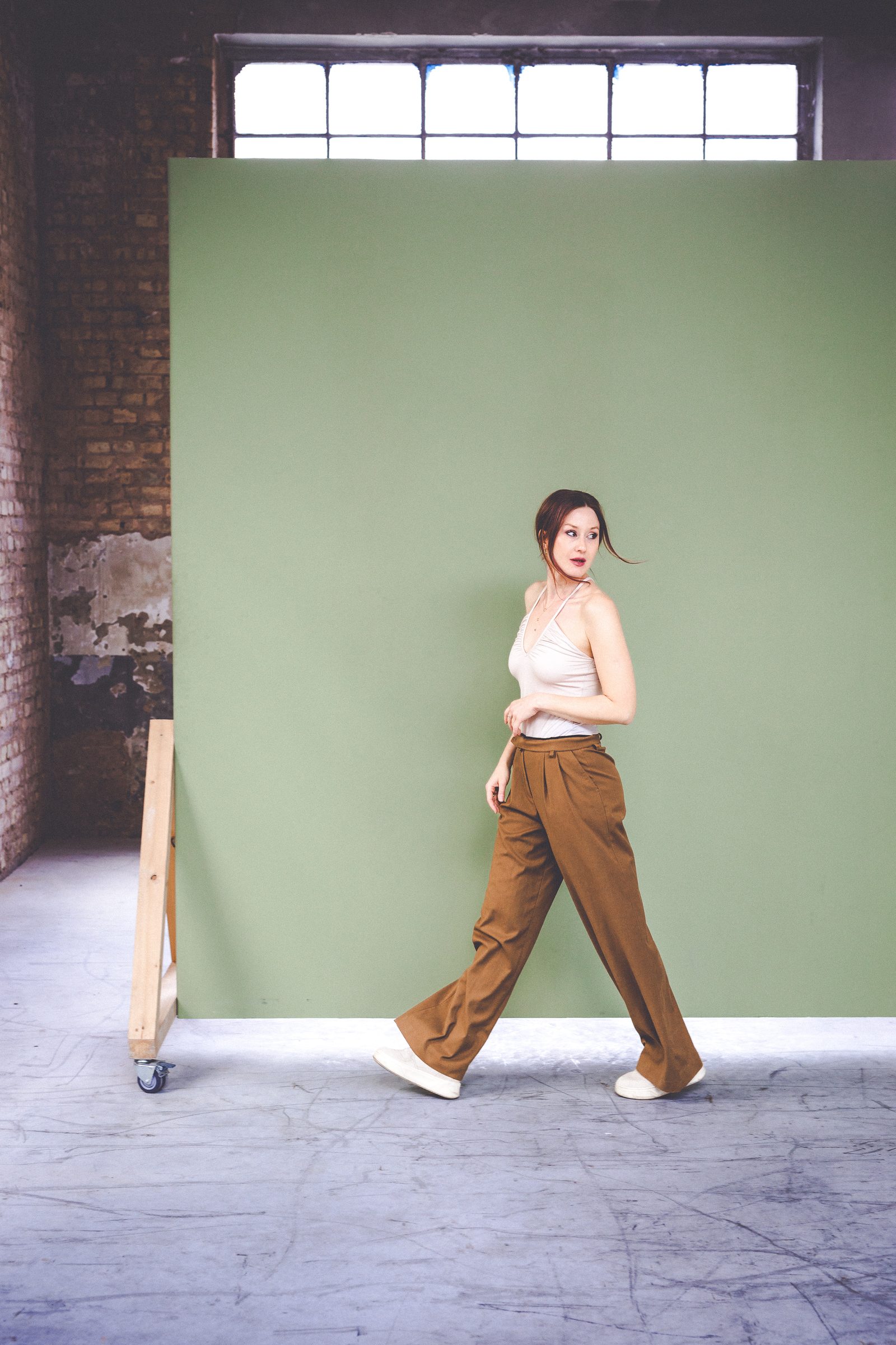 Sasha Walker geht in weißem Top und weiter brauner Hose vor einem grünen Fotohintergrund durch das Loft
