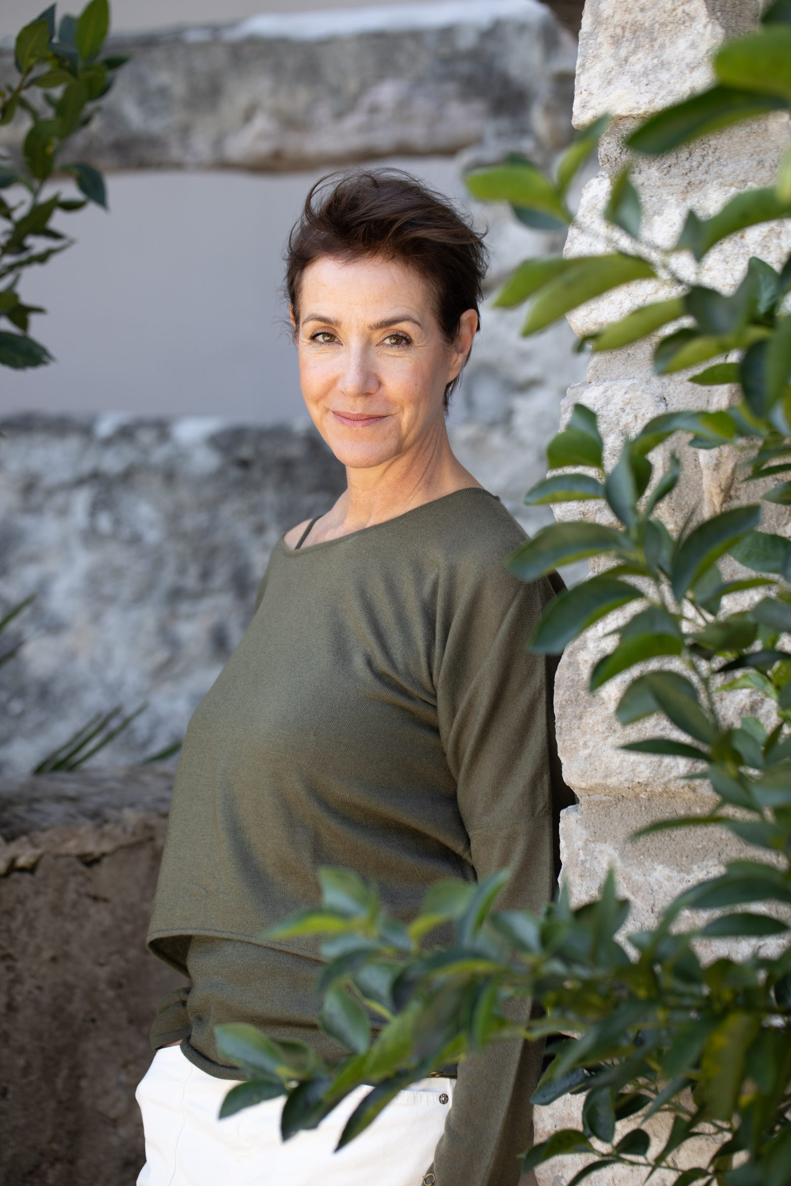 Frau in olivgrünem Shirt lehnt lächelnd an einer Natursteinmauer zwischen grünen Blättern, Personal-Branding-Porträt für Schminktante
