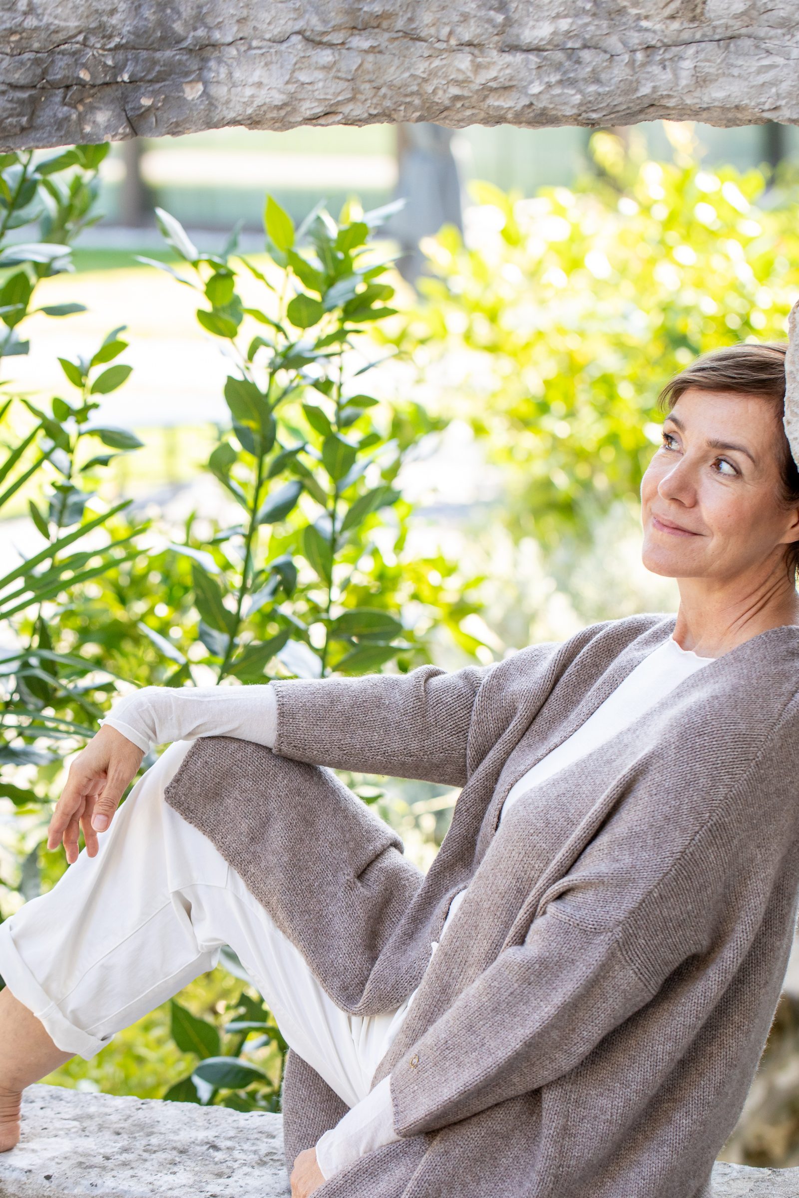 Frau in grauer Strickjacke sitzt entspannt vor sonnigen grünen Büschen und schaut zur Seite, Personal-Branding-Porträt für Schminktante