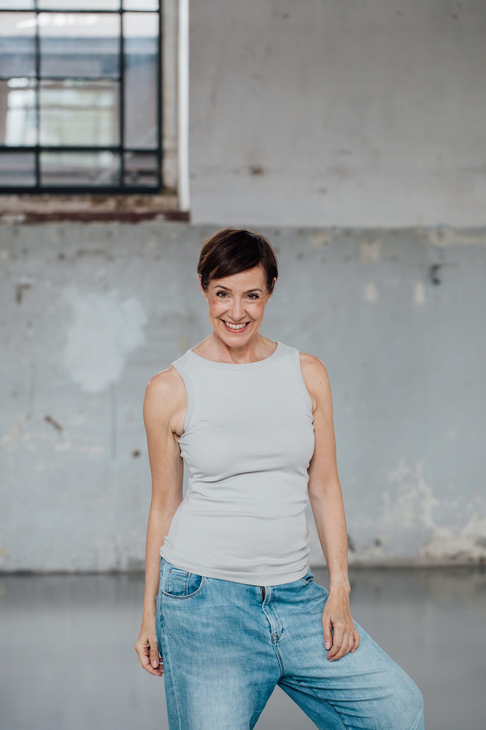 Frau in hellgrauem Top und Jeans steht lachend vor einer grauen Betonwand in einer Industriehalle, Personal-Branding-Porträt für Schminktante