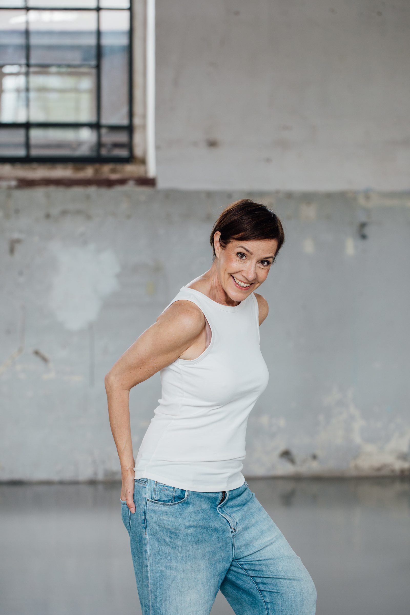 Frau in hellem Top und Jeans posiert mit der Hand an der Hüfte vor einer grauen Betonwand, Personal-Branding-Porträt für Schminktante