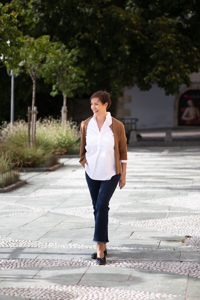 Frau in weißer Bluse, brauner Jacke und dunkler Hose geht lächelnd über einen gepflasterten Innenhof mit Bäumen, Personal-Branding-Porträt für Schminktante