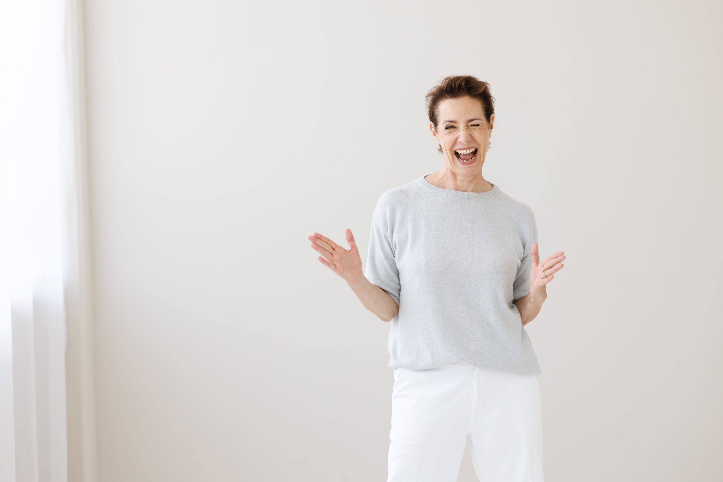 Lachende Frau mit kurzen Haaren in hellem Shirt vor einer hellen Wand