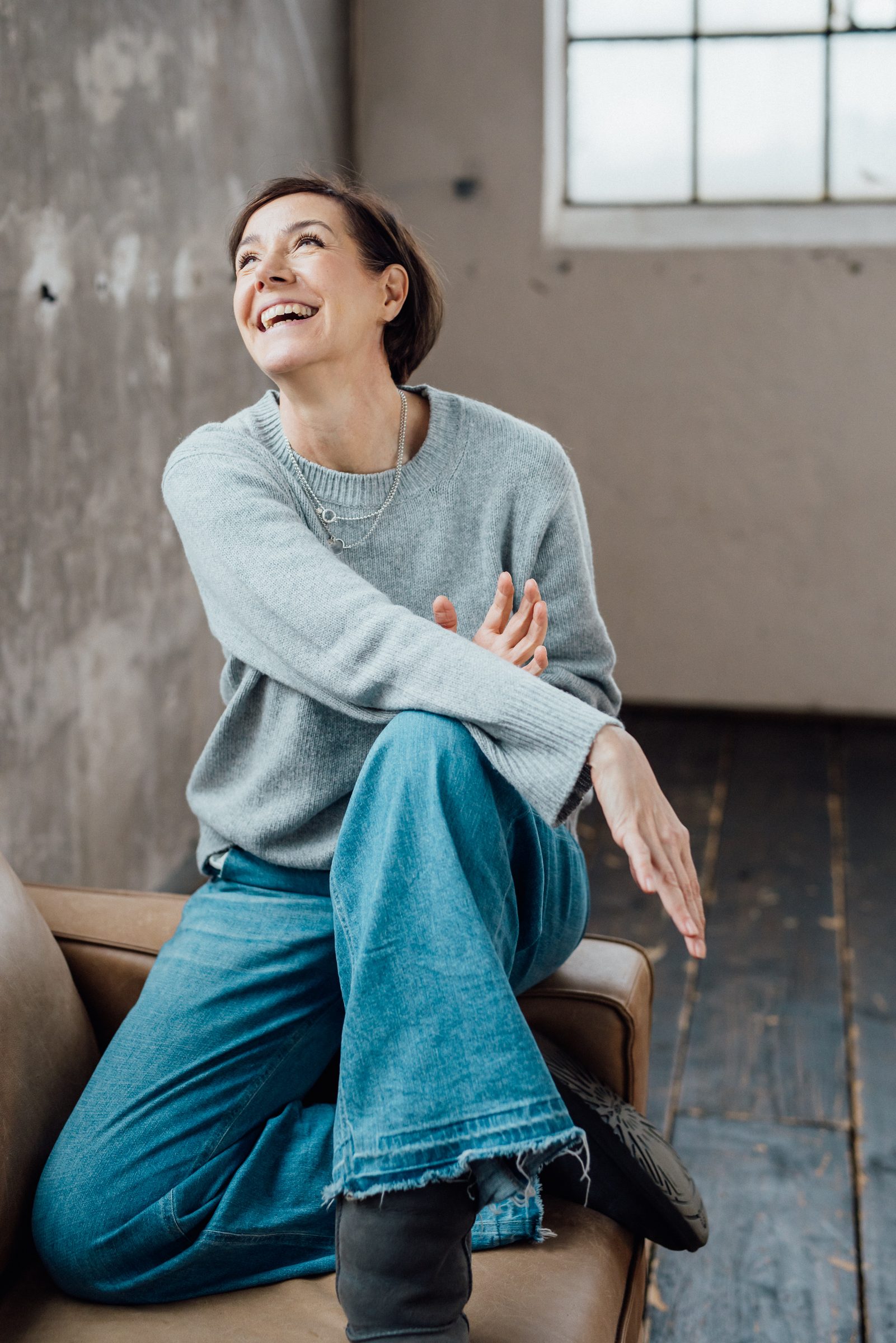 Frau in hellblauem Pullover und weiter Jeans sitzt lachend auf einem Sessel in einem Loft, Personal-Branding-Porträt für Schminktante