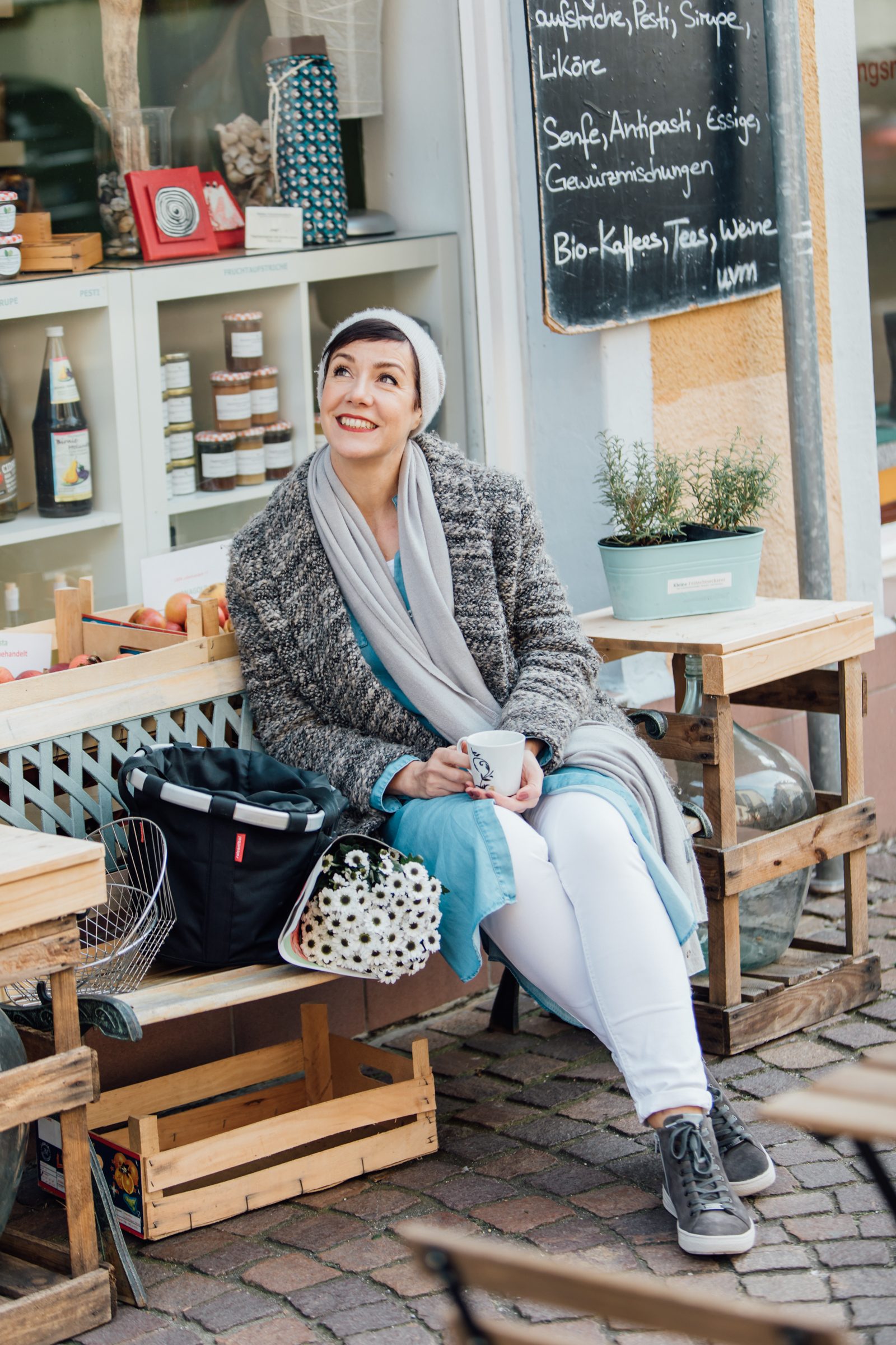 Frau mit grauem Schal und Strickjacke sitzt mit einer Tasse vor einem kleinen Laden mit Regalen und Kreidetafel, Personal-Branding-Porträt für Schminktante