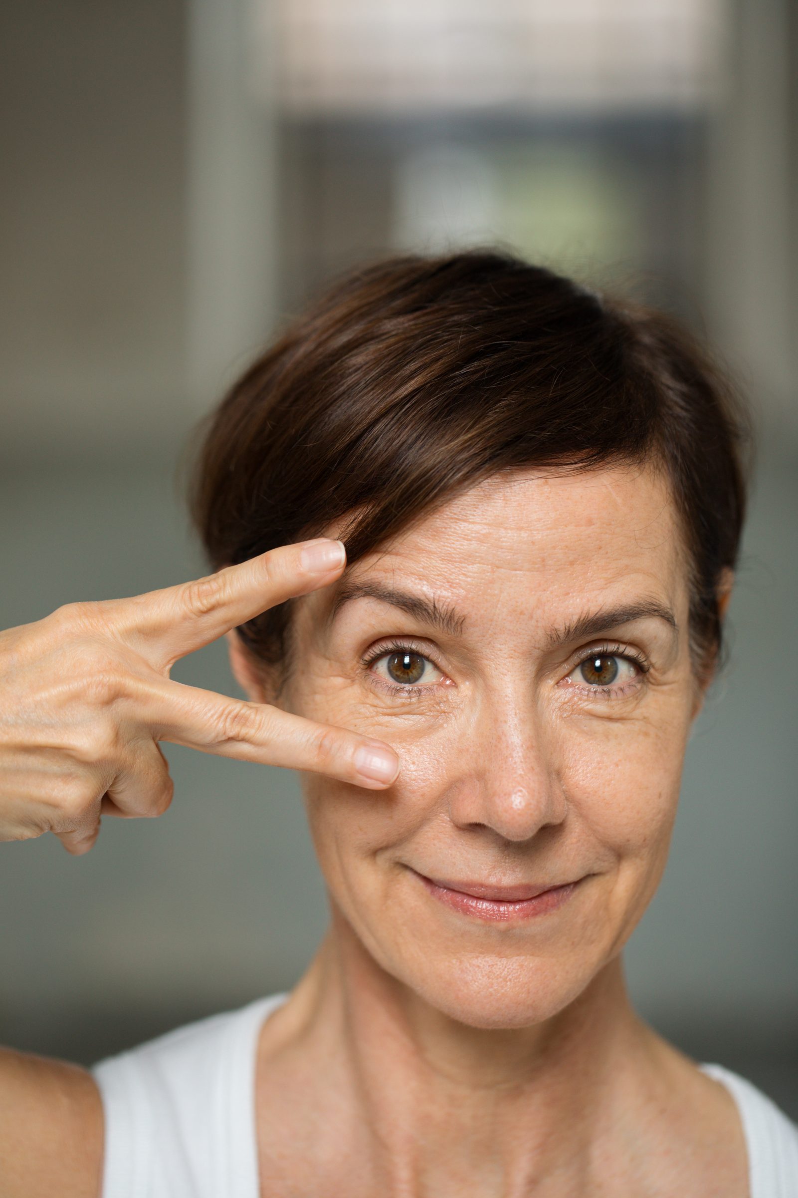Nahaufnahme einer lächelnden Frau mit kurzen dunklen Haaren, die zwei Finger neben ihr Auge hält, Personal-Branding-Porträt für Schminktante