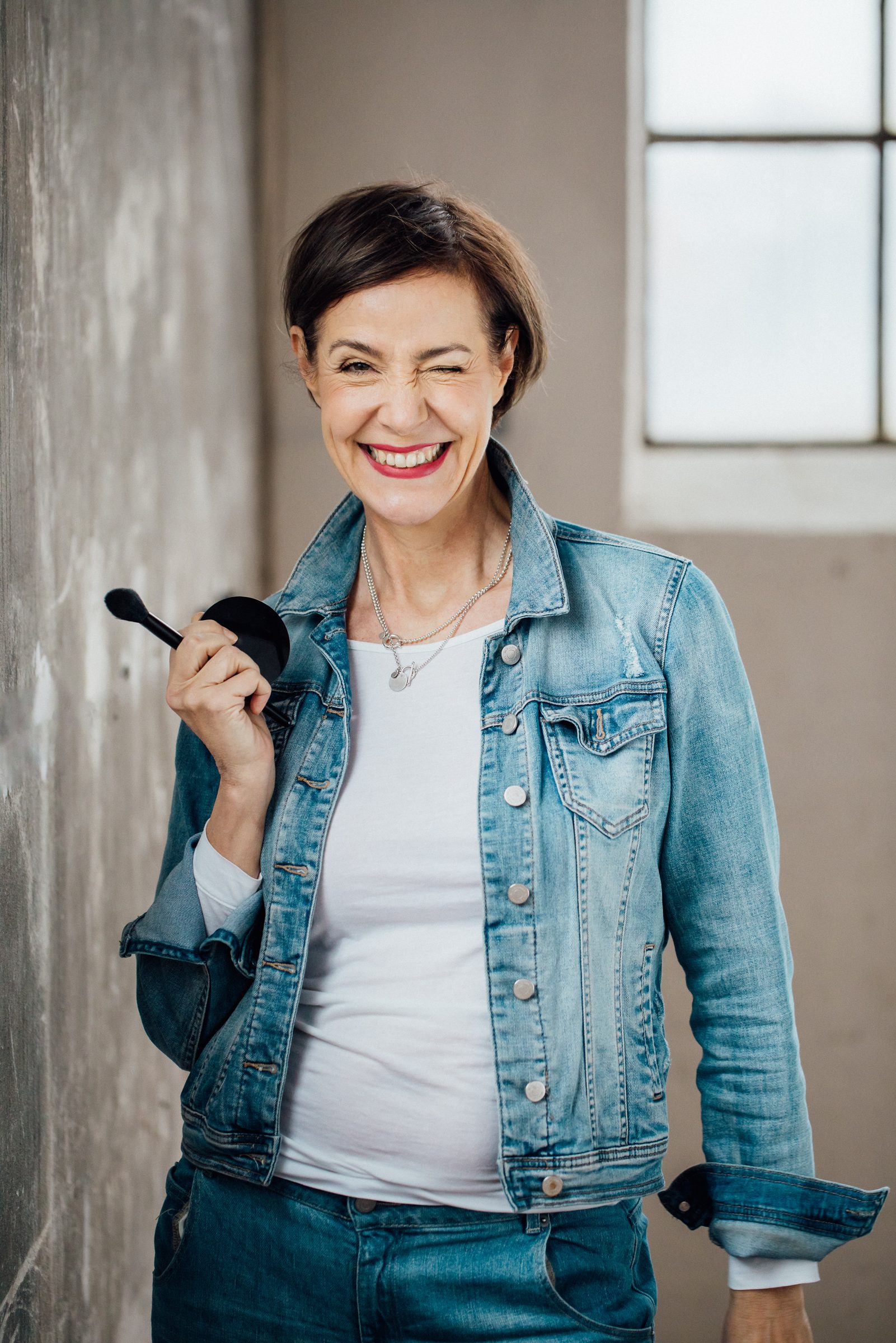Frau in Jeansjacke zwinkert lachend und hält einen Make-up-Pinsel und eine Puderdose, Personal-Branding-Porträt für Schminktante