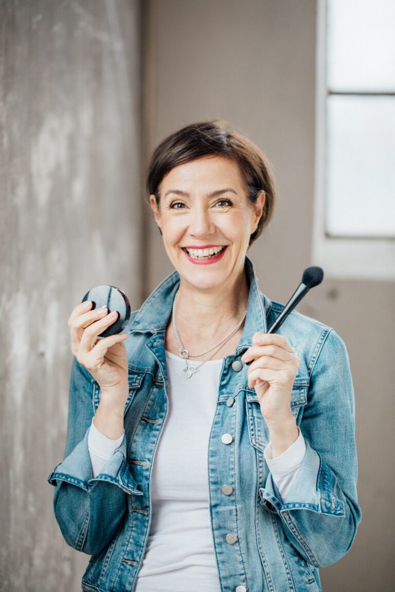 Lachende Frau in Jeansjacke mit Puderdose und Pinsel, Personal-Branding-Porträt für Schminktante