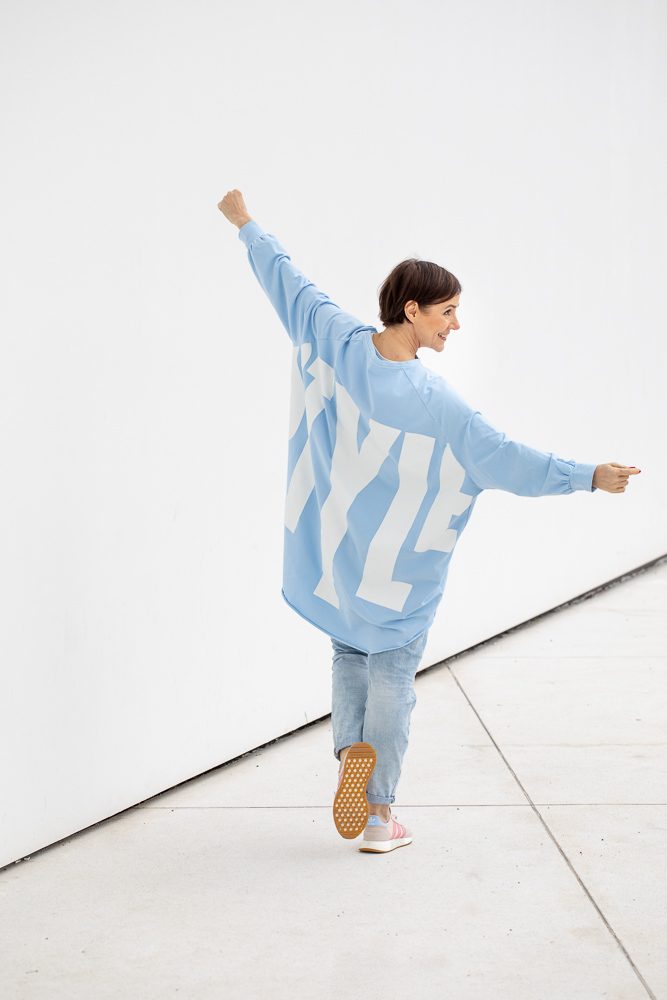 Frau in weitem hellblauem Pullover mit Schriftzug streckt tanzend die Arme vor einer weißen Wand aus, Personal-Branding-Porträt für Schminktante