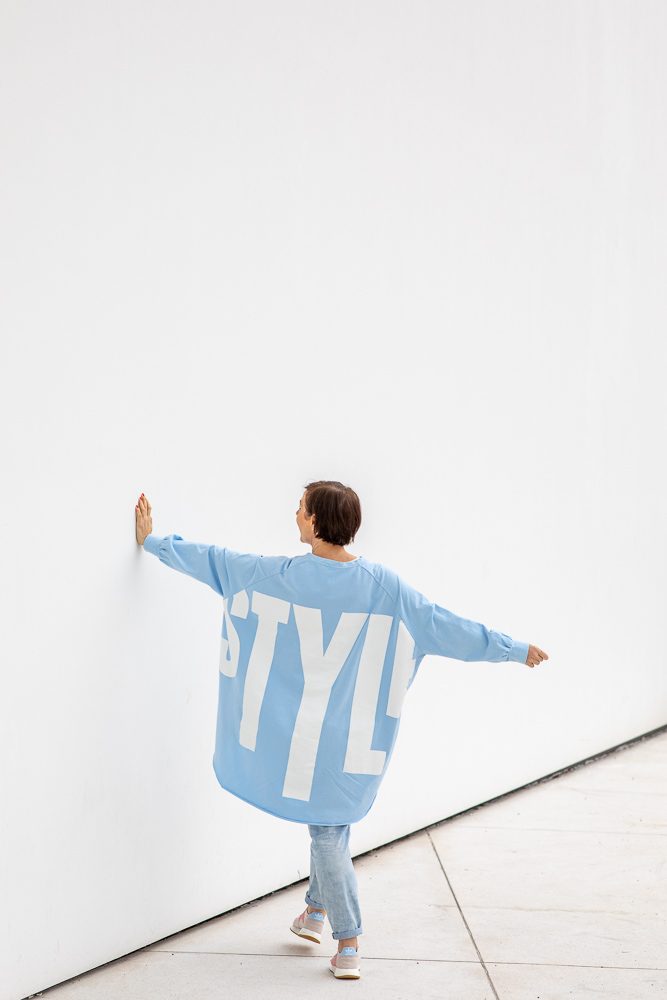 Frau in weitem hellblauem Pullover mit Schriftzug stützt sich mit ausgestreckten Armen an einer weißen Wand ab, Rückenansicht, Personal-Branding-Porträt für Schminktante