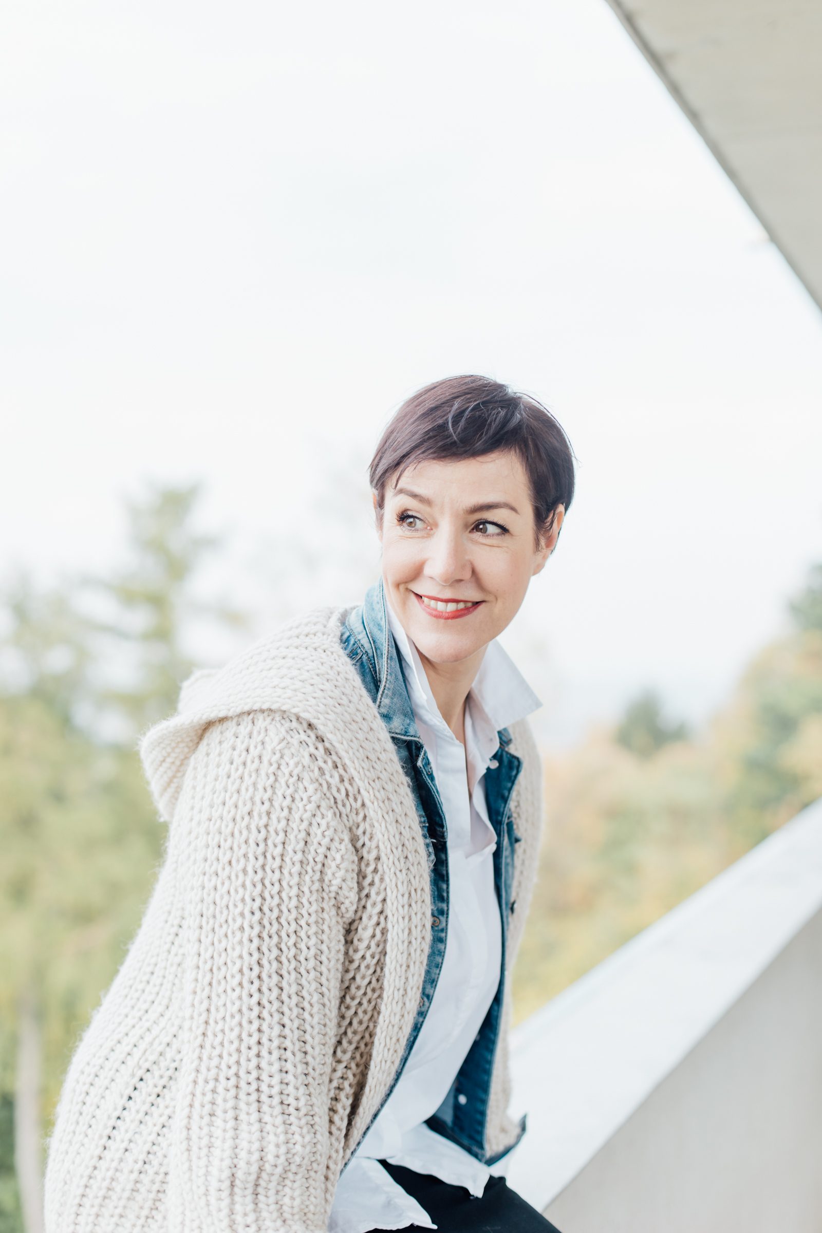 Frau mit kurzen dunklen Haaren in heller Strickjacke und Jeanshemd schaut lächelnd zur Seite, im Hintergrund Bäume, Personal-Branding-Porträt für Schminktante