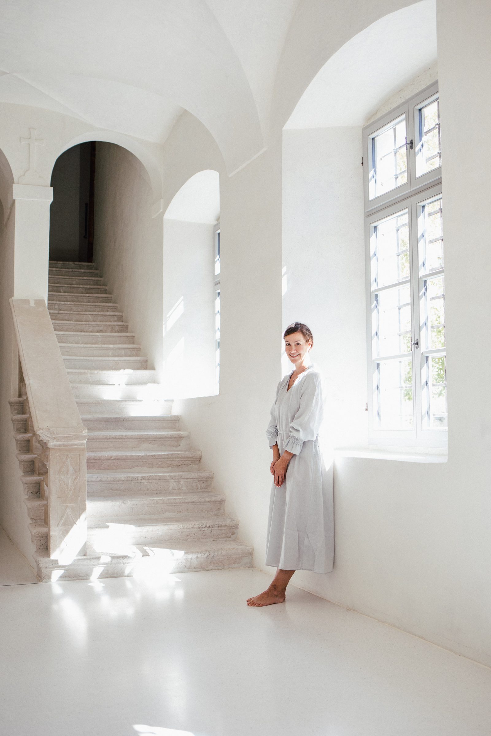 Frau in hellem Kleid steht barfuß am Fenster eines weißen Treppenhauses mit Steintreppe und Rundbögen, Personal-Branding-Porträt für Schminktante
