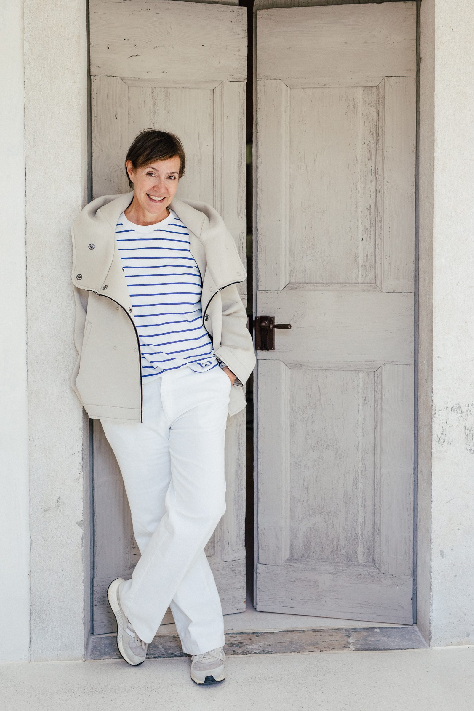 Frau in gestreiftem Shirt, heller Jacke und weißer Hose lehnt lächelnd an einer alten grauen Holztür, Personal-Branding-Porträt für Schminktante