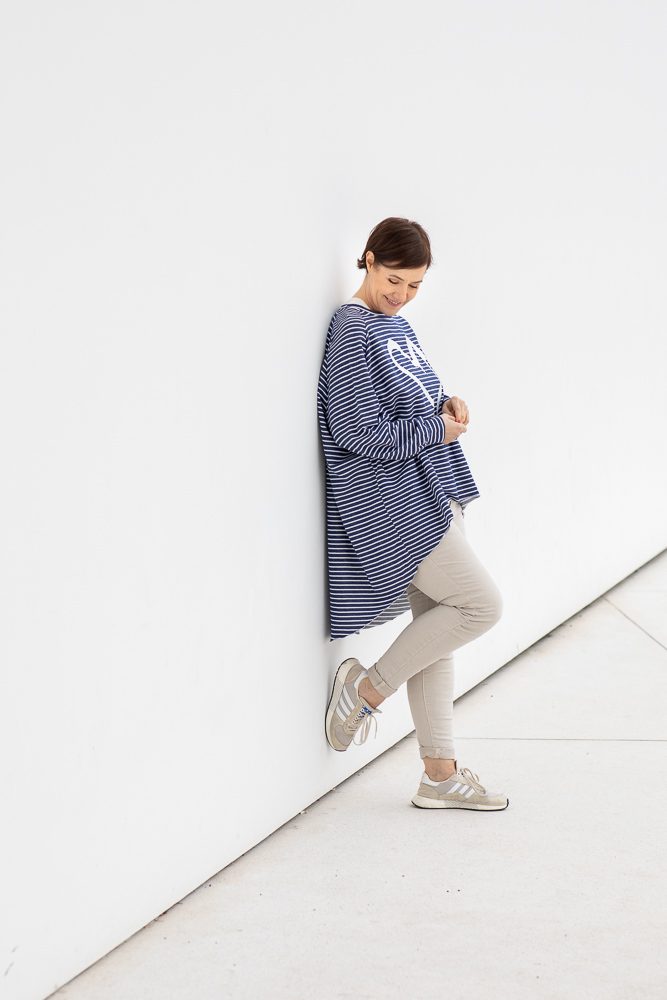Frau in gestreiftem Longpullover lehnt lächelnd an einer weißen Wand, Personal-Branding-Porträt für Schminktante
