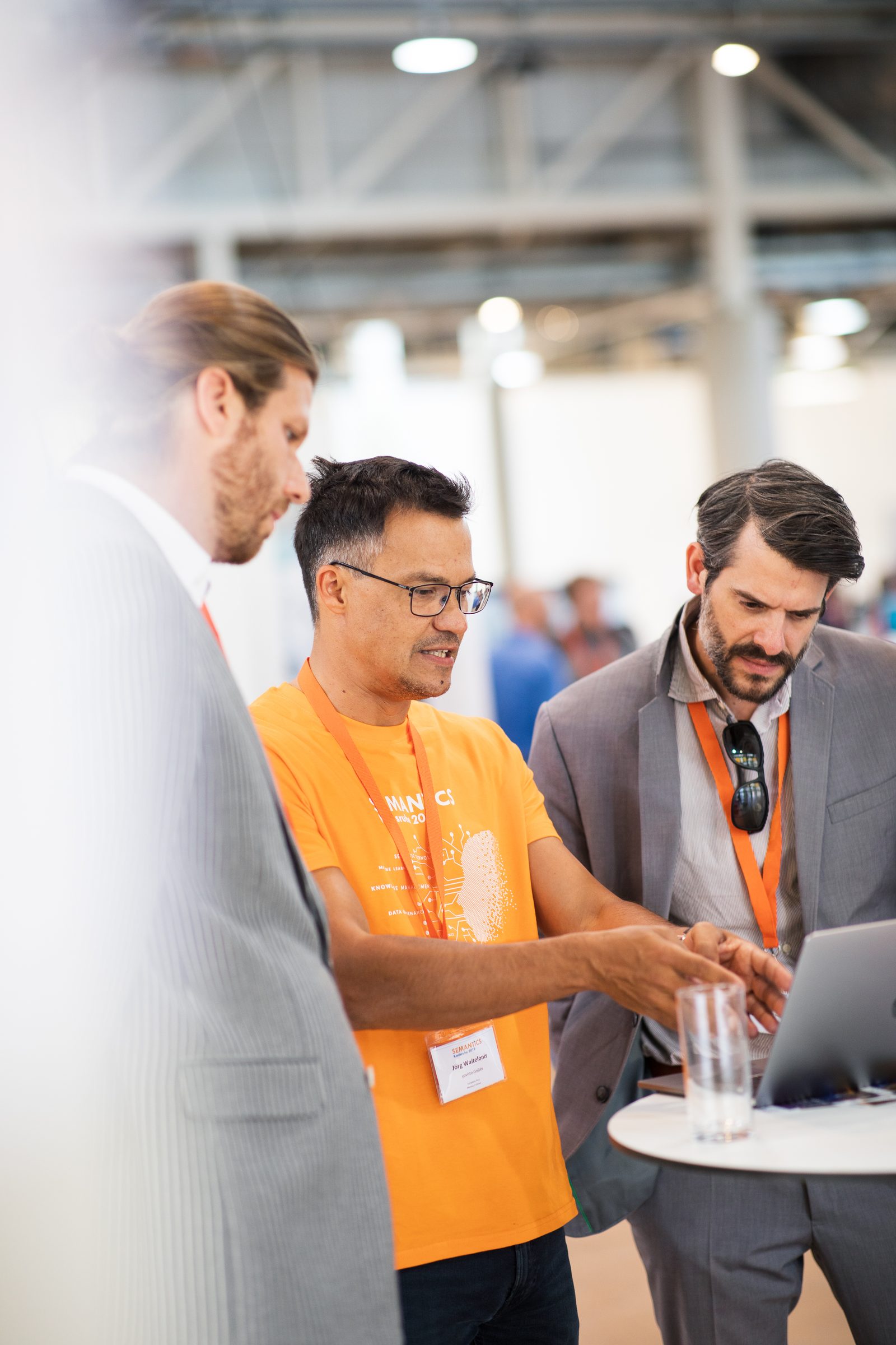 Drei Männer schauen am Stehtisch gemeinsam auf einen Laptop, einer im orangen Shirt der Konferenz SEMANTiCS erklärt etwas