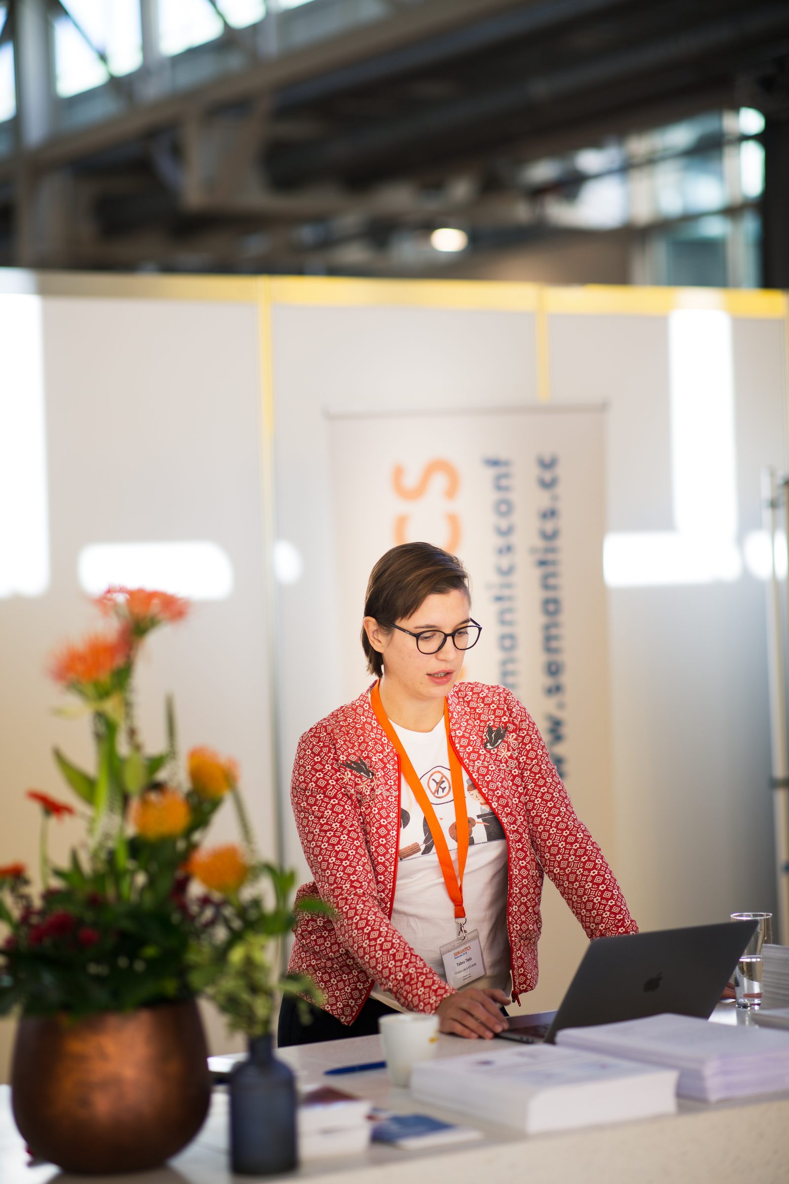 Frau mit Brille und roter Jacke am Empfangstresen der Konferenz SEMANTiCS, vor ihr ein Laptop und ein Blumenstrauß