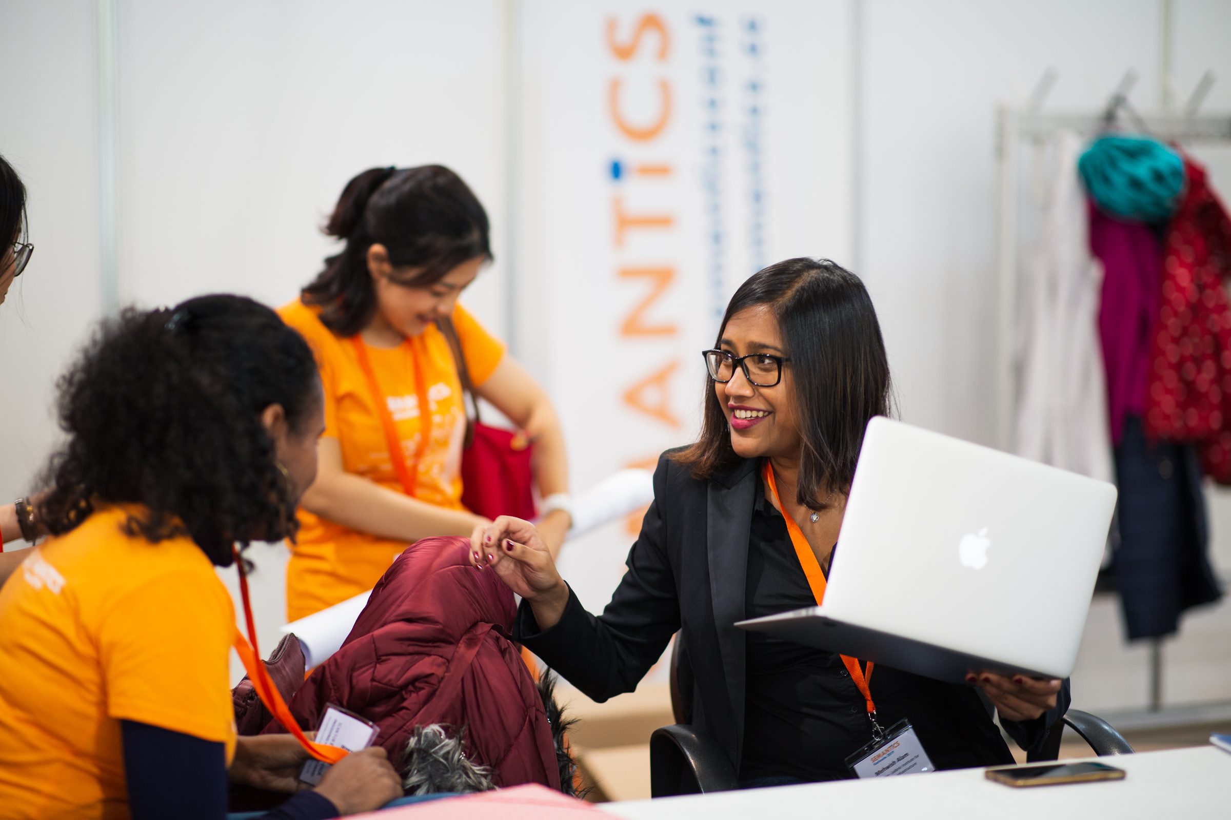 Lächelnde Frau mit Laptop an der Registrierung der Konferenz SEMANTiCS, daneben Helferinnen in orangen Team-Shirts