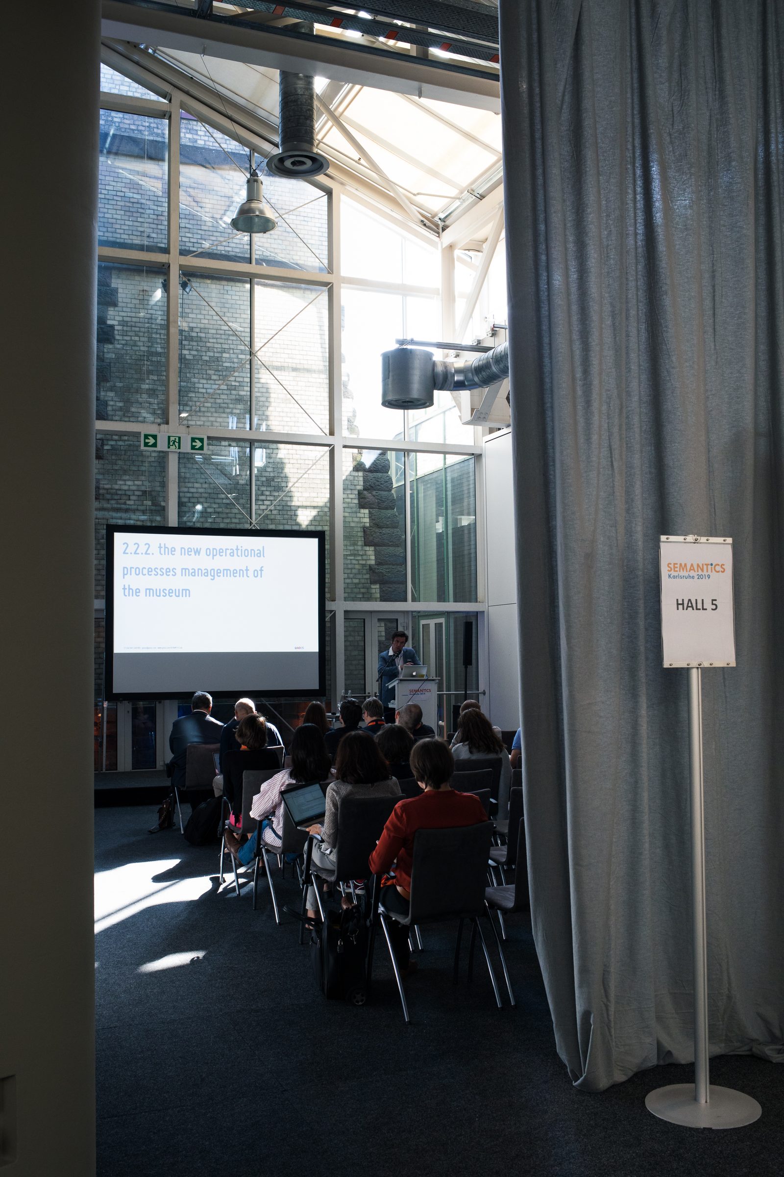 Blick an einem Vorhang vorbei in einen hellen Vortragssaal der Konferenz SEMANTiCS mit Publikum und Leinwand