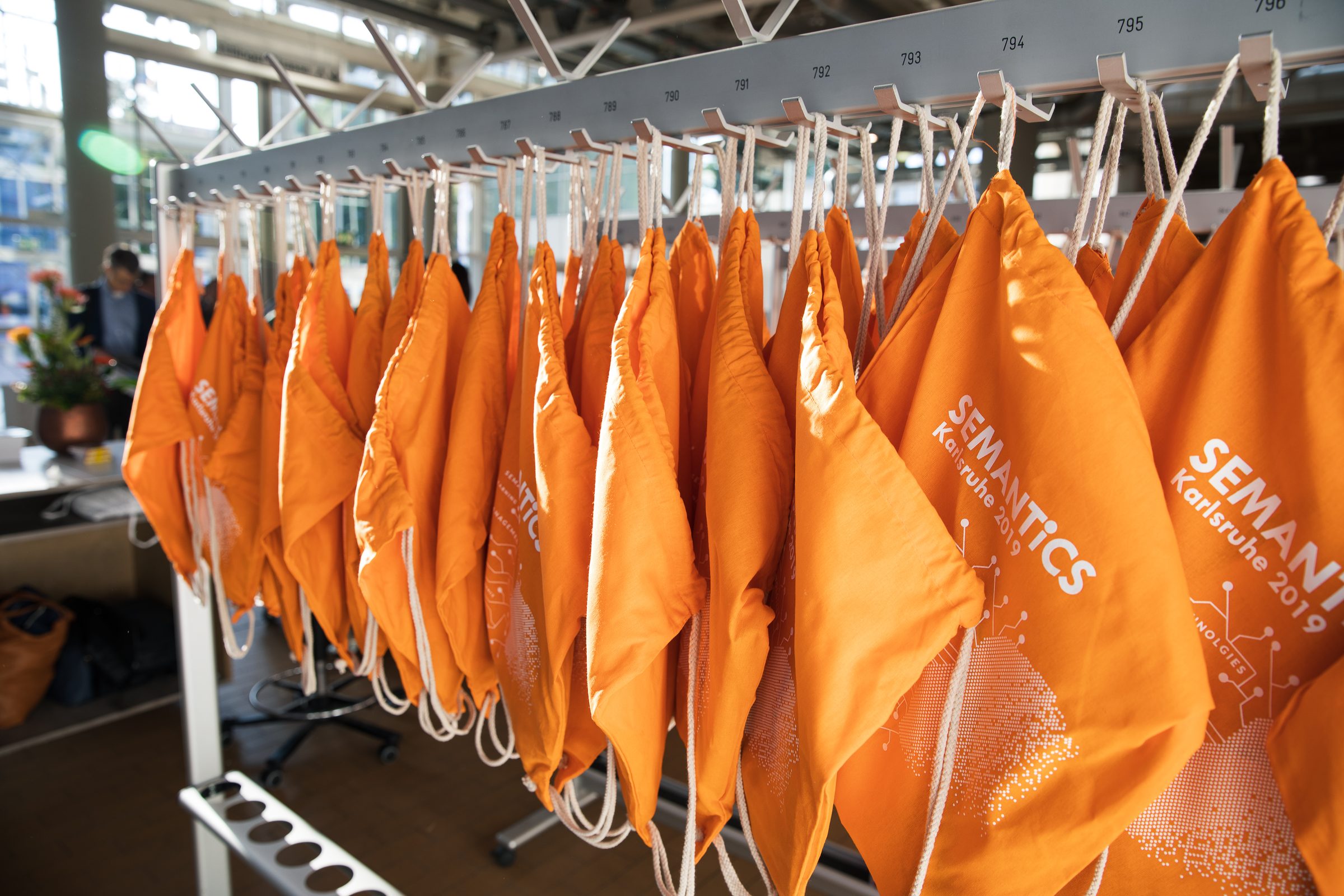 Orange Turnbeutel mit dem Aufdruck SEMANTiCS Karlsruhe 2019 hängen nebeneinander an einer Garderobe