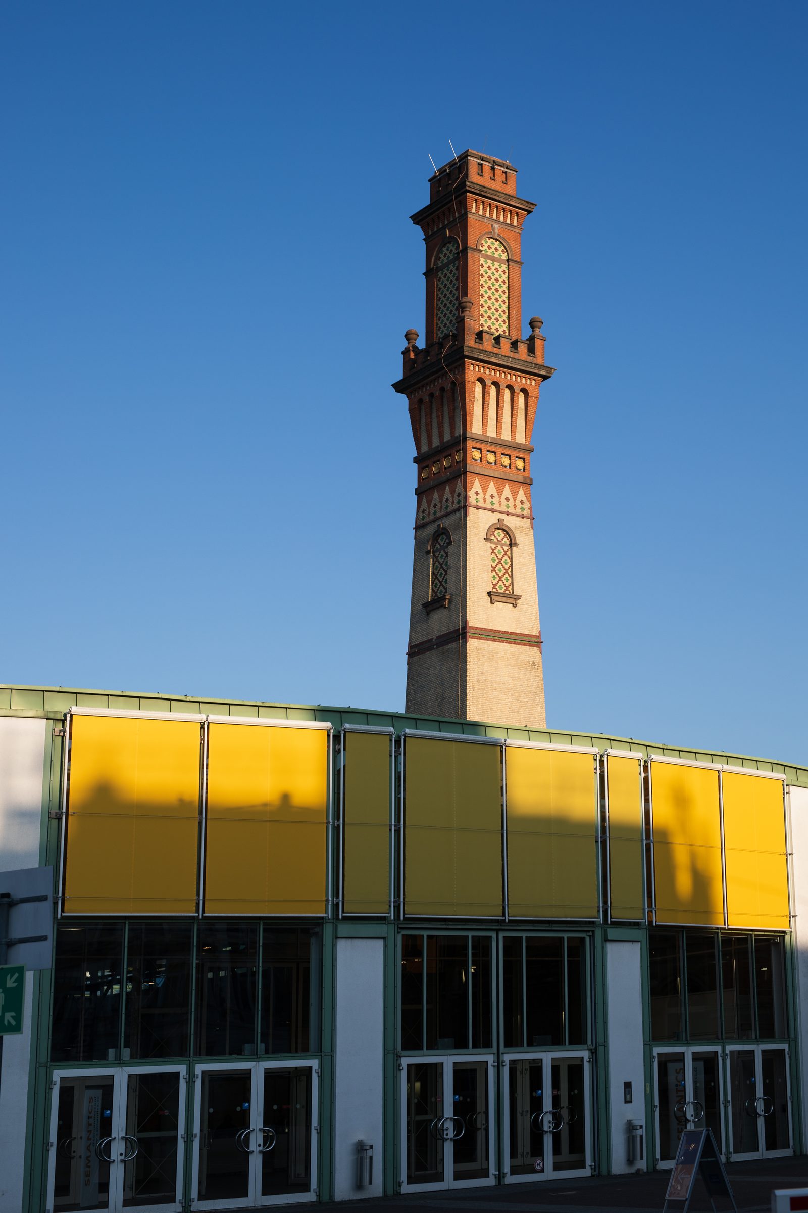 Historischer Backsteinturm vor blauem Himmel über einer Halle mit gelber Fassade am Veranstaltungsort der Konferenz SEMANTiCS in Karlsruhe