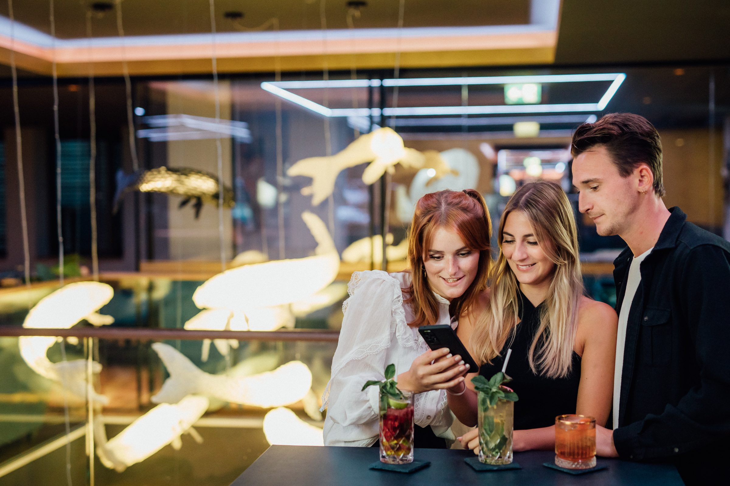 Drei junge Leute schauen abends an einem Bartisch mit Cocktails gemeinsam auf ein Smartphone, Kampagne der Stadtwerke Heilbronn