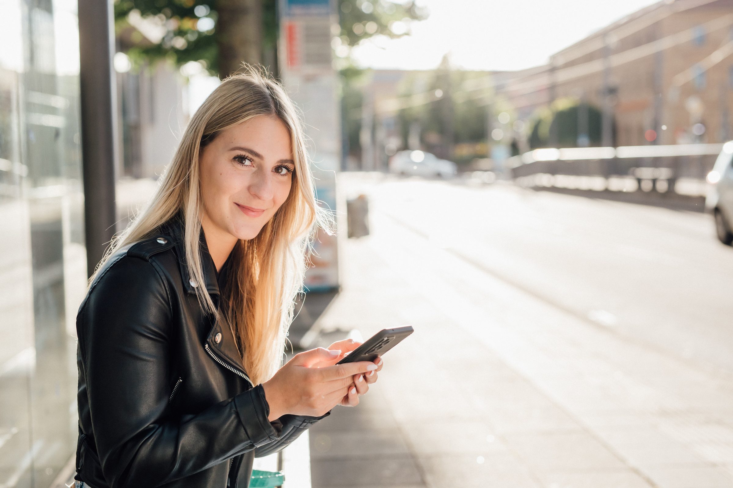 Junge Frau mit Smartphone in der Hand steht an einer sonnigen Straße und lächelt in die Kamera, Kampagne der Stadtwerke Heilbronn