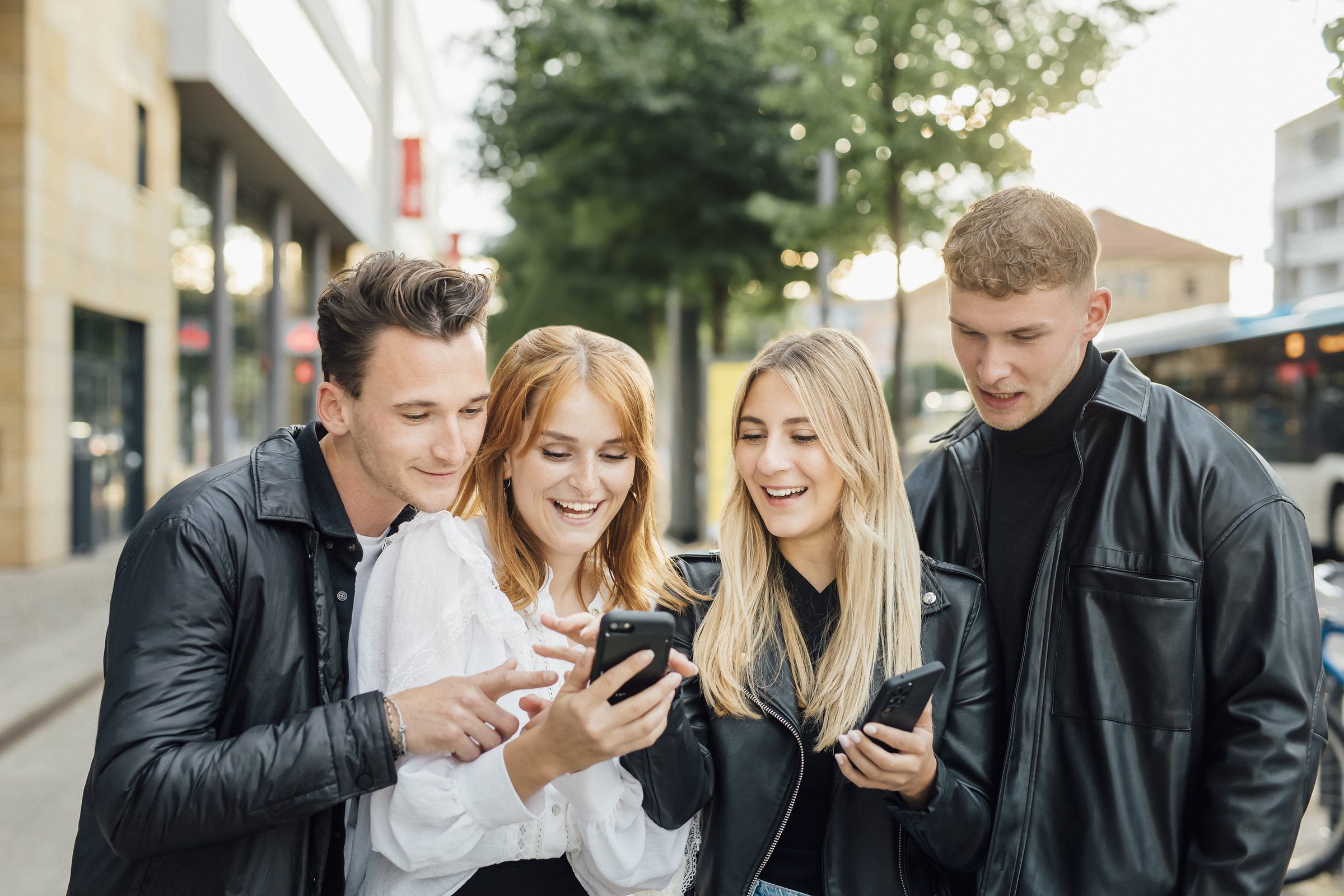 Vier junge Leute schauen lachend gemeinsam auf ein Smartphone, im Hintergrund ein Bus, Kampagne der Stadtwerke Heilbronn