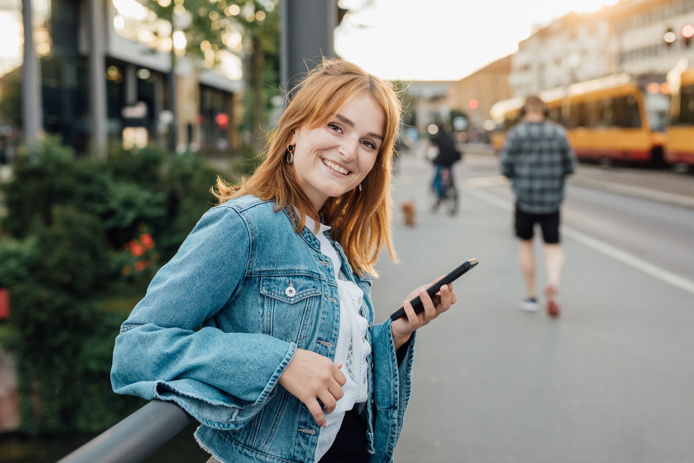 Junge Frau in Jeansjacke lehnt lächelnd mit dem Smartphone an einem Geländer, im Hintergrund eine Stadtbahn, Kampagne der Stadtwerke Heilbronn