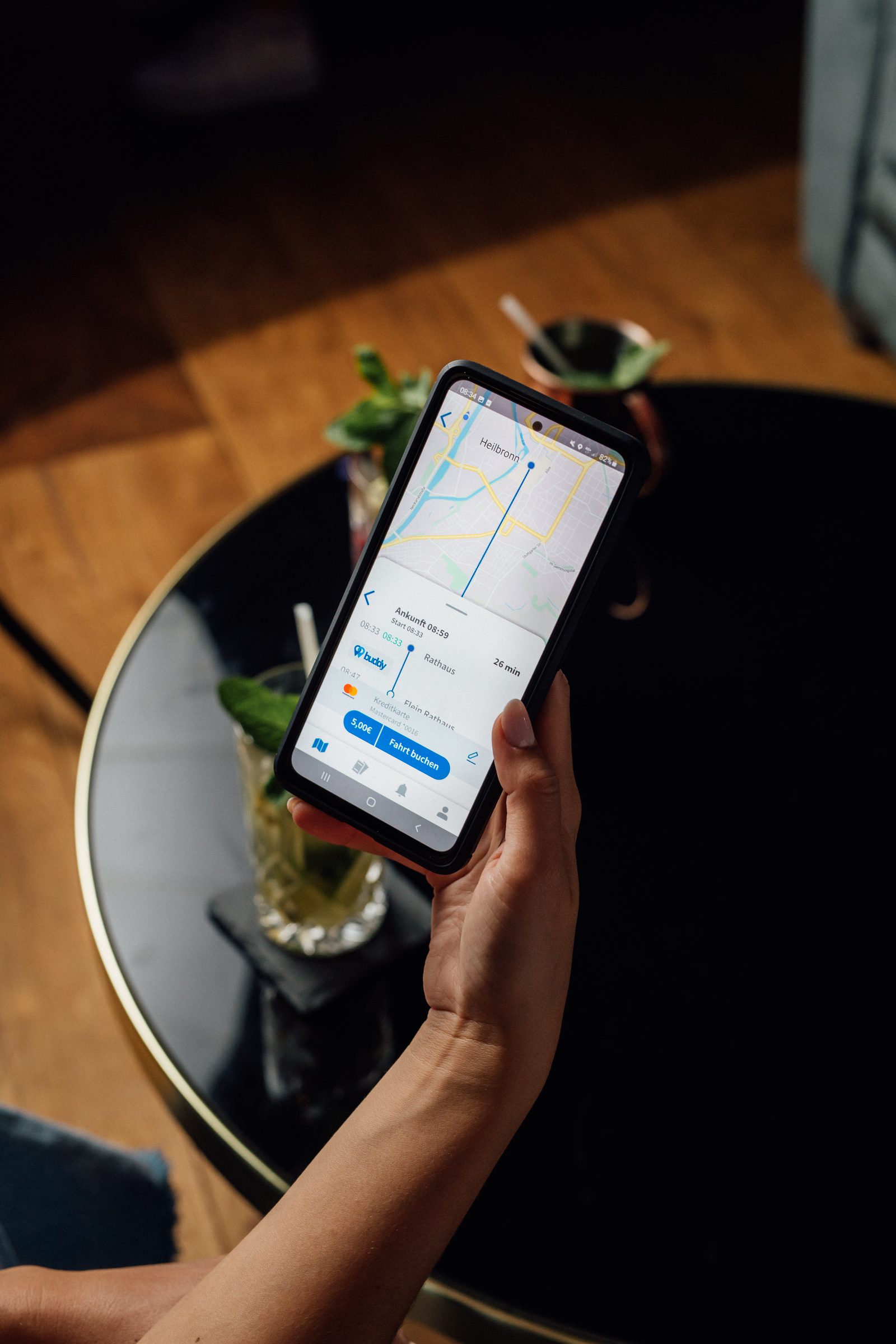 Hand hält ein Smartphone mit der Buchungsseite des Nachtshuttles buddy über einem Bartisch mit Cocktail, Kampagne der Stadtwerke Heilbronn