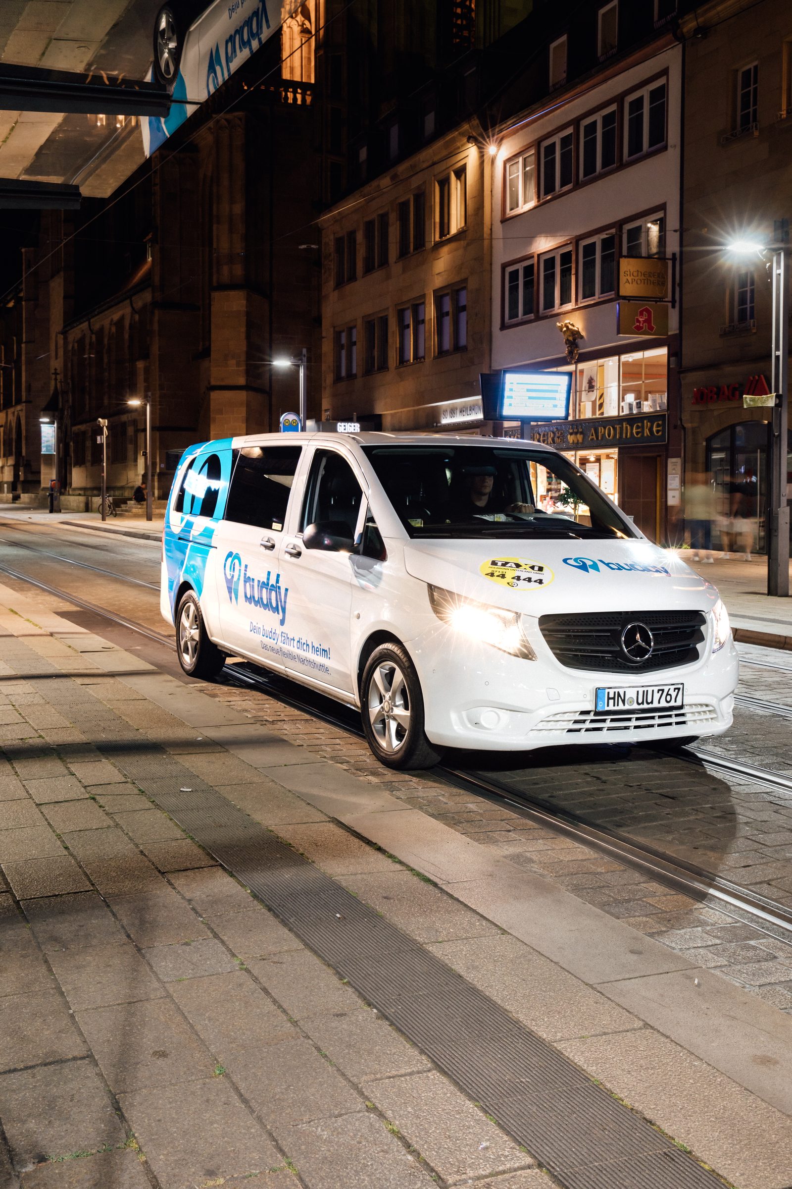 Weißer Kleinbus des Nachtshuttles buddy fährt nachts mit eingeschaltetem Licht durch eine beleuchtete Innenstadtstraße, Kampagne der Stadtwerke Heilbronn