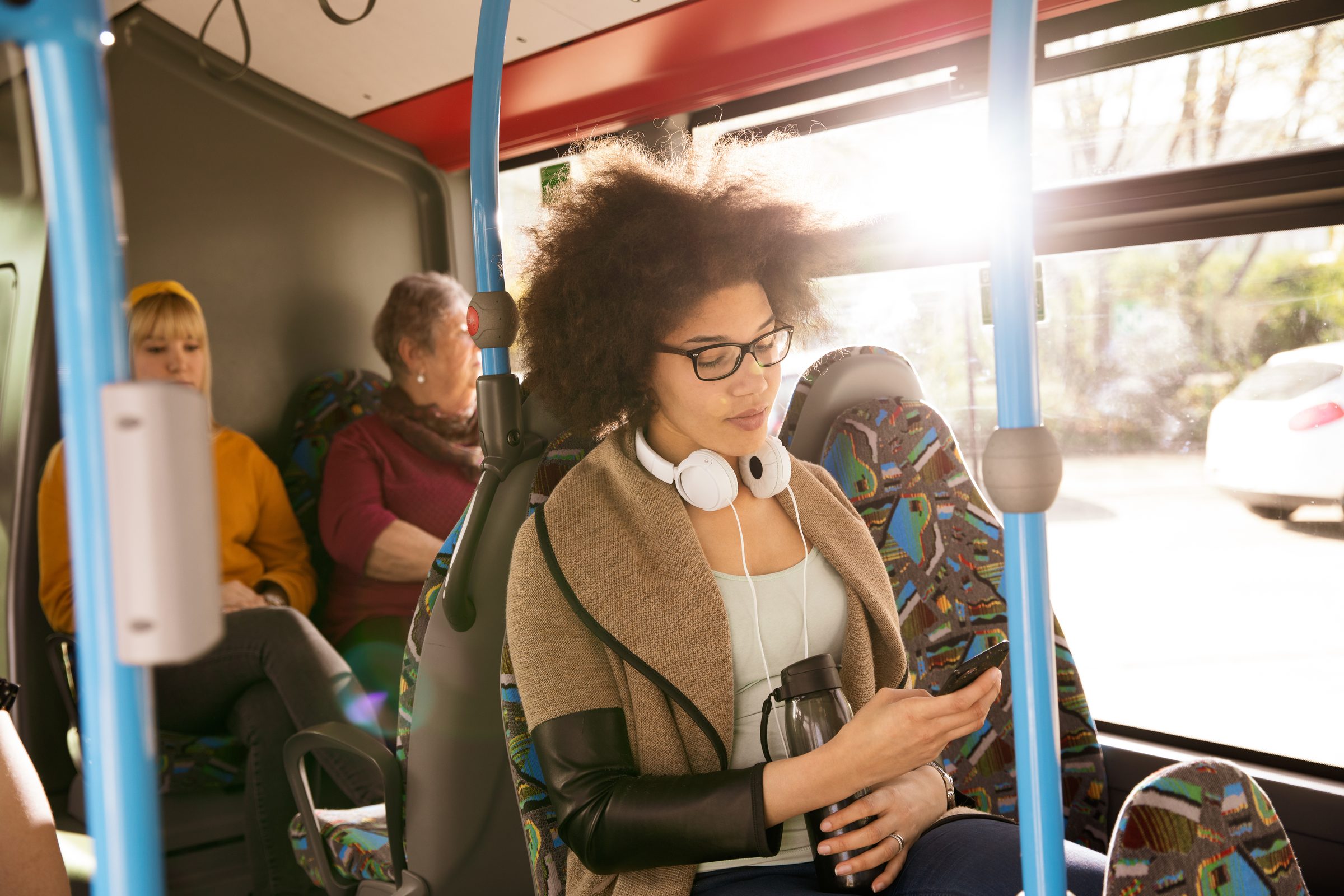 Junge Frau mit Kopfhörern um den Hals schaut im Bus auf ihr Smartphone, hinter ihr weitere Fahrgäste, Stadtwerke Heilbronn