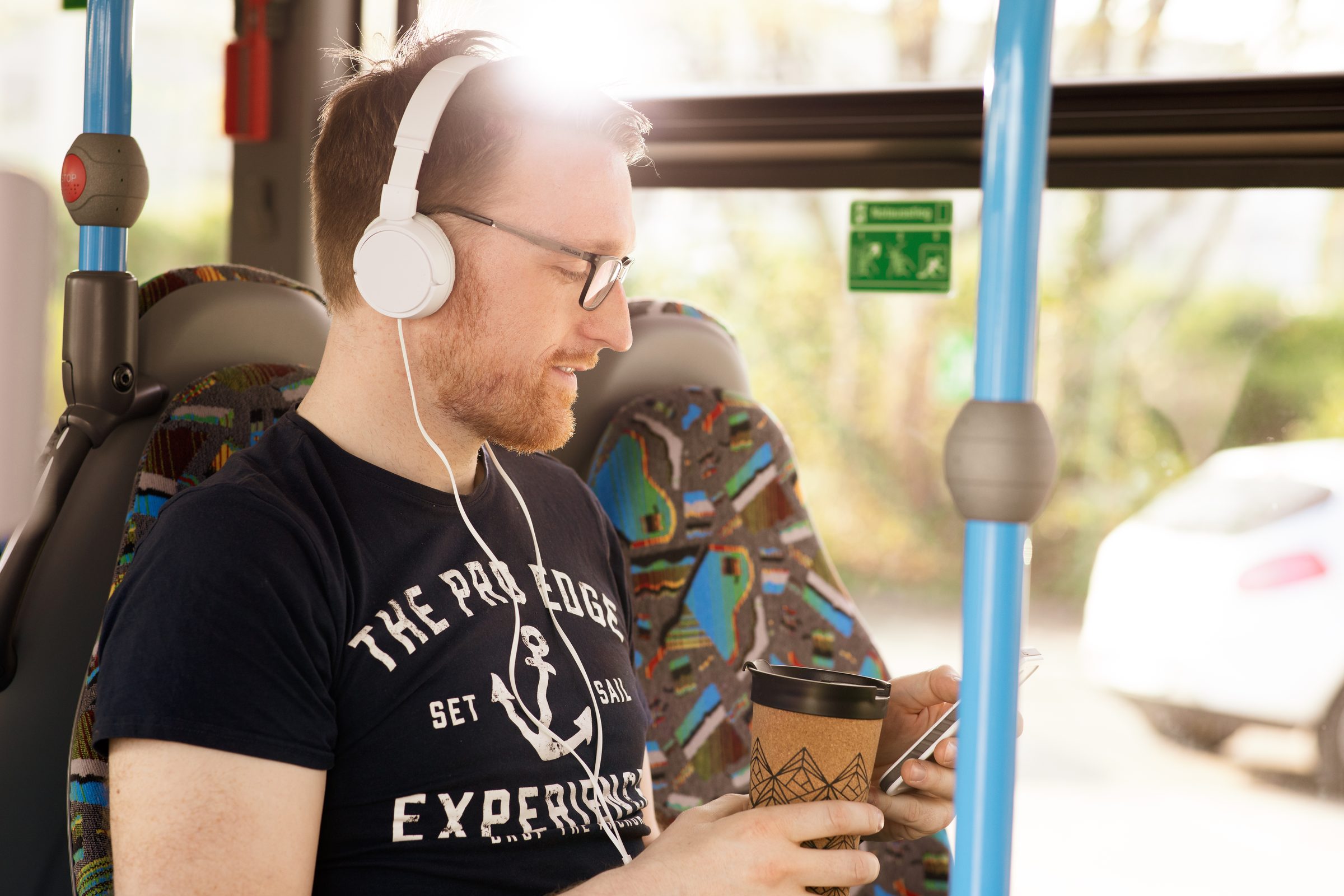 Mann mit weißen Kopfhörern und Kaffeebecher sitzt lächelnd im Bus und schaut auf sein Smartphone, Stadtwerke Heilbronn