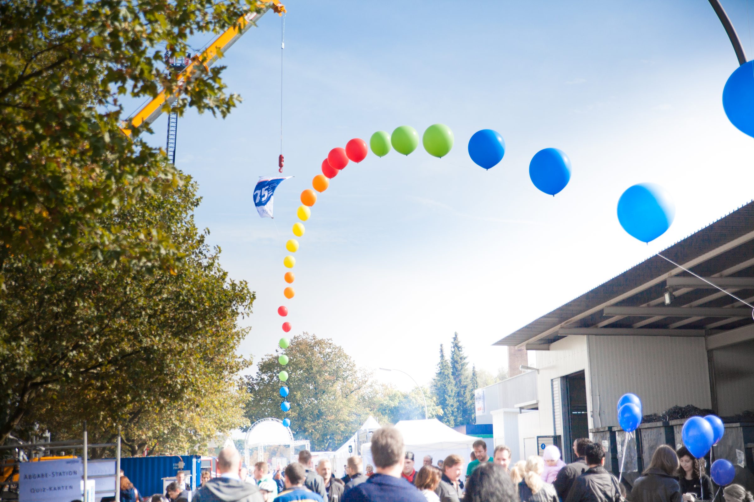 Bunte Ballonkette an einem Kran über dem Werksgelände von Weber Hydraulik, darunter Besucherinnen und Besucher beim Firmenjubiläum